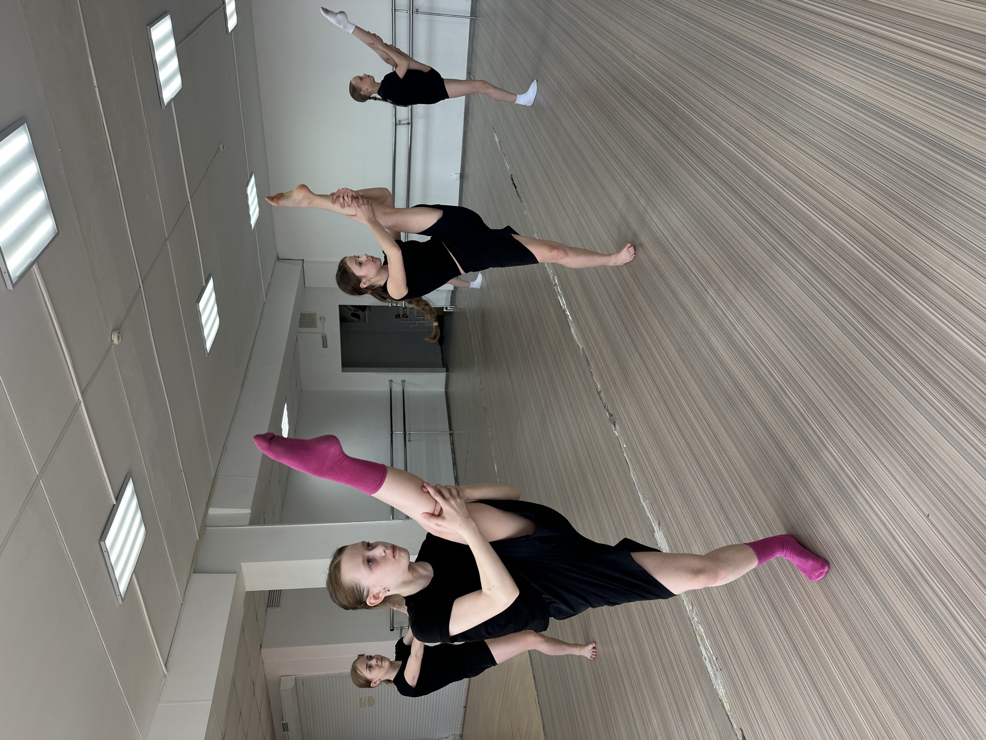 The image shows four young women stretching in a dance studio. The studio has a light-colored wooden floor and white walls. There are fluorescent lights on the ceiling. All four women are wearing black shirts and shorts. One woman has a pink sock on her extended leg. Another woman has a white sock on her leg. They are all stretching their legs, holding them with their hands. The location is a dance studio, likely in Votkinsk, Russia.