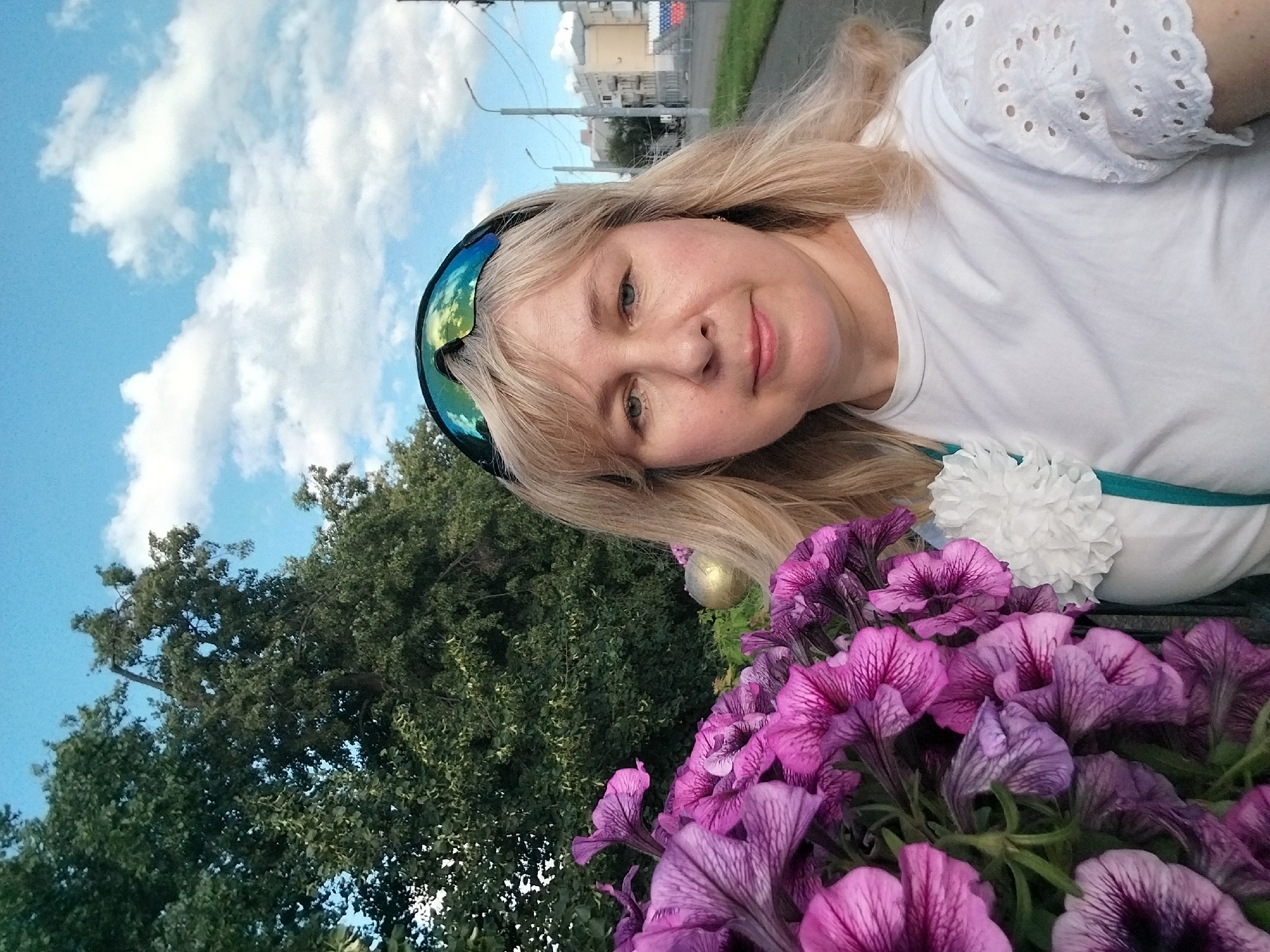 A medium shot shows a woman with blonde hair and blue eyes wearing a white t-shirt and sunglasses. She is outdoors, with purple flowers in the foreground and trees and buildings in the background. The sky is blue with white clouds. The location is the city of Orenburg, Russia.