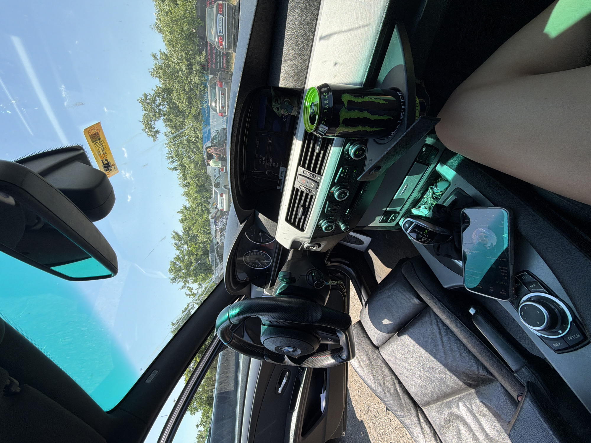 The image is an interior view of a car, likely a BMW, parked on a street in Burgas, Bulgaria. The dashboard, steering wheel, and leather seats are visible. A can of Monster energy drink and a smartphone are placed on the center console. In the background, other cars and trees are seen, suggesting an outdoor parking area. The sky is clear and blue, indicating a sunny day. A Bulgarian vignette is attached to the windshield. The overall scene depicts a casual setting inside a parked vehicle on a summer day.
