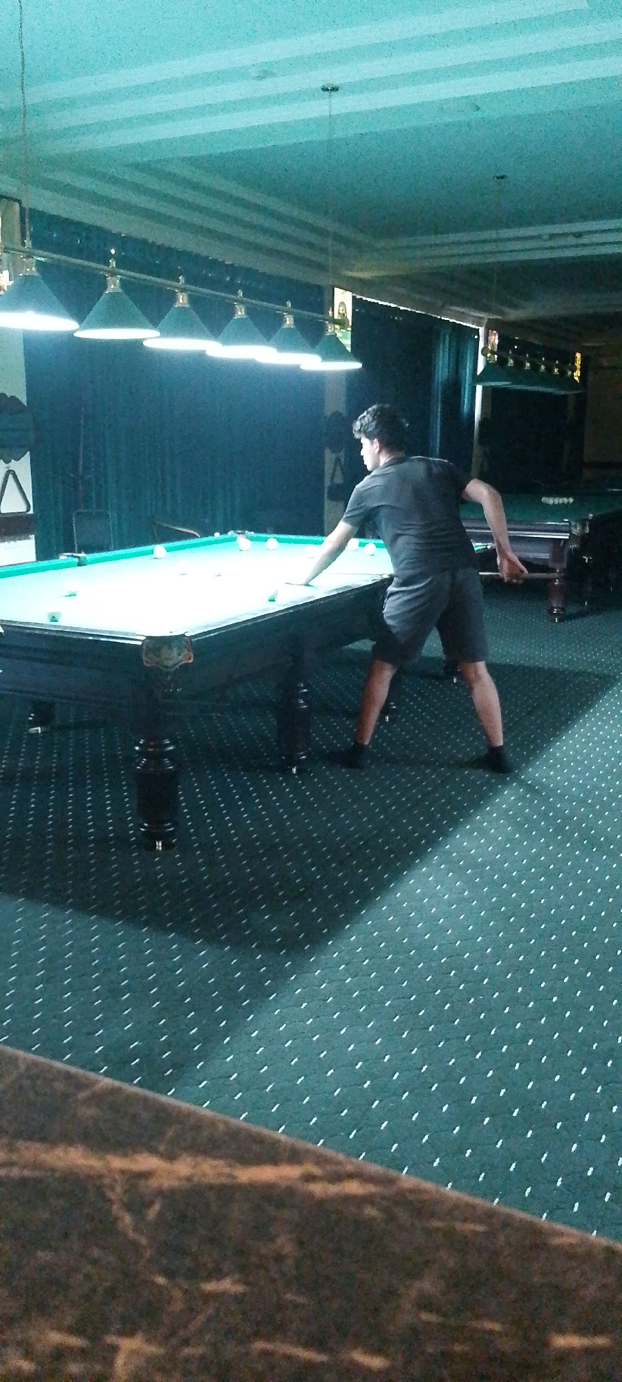 A young man wearing a dark t-shirt and dark shorts is playing billiards in a dimly lit room. He is positioned behind a pool table, holding a cue stick and appearing to be in the process of taking a shot. The pool table has a green felt surface and numerous white balls scattered across it. Above the table, a row of green lamps hangs, illuminating the playing area. The room has teal-colored walls and a patterned teal carpet on the floor. In the background, there are dark green curtains and other pool tables. The lighting casts long shadows on the carpet, creating a somewhat moody atmosphere. The overall impression is of a recreational setting, likely a billiard hall, located in the city of Angren, Uzbekistan.