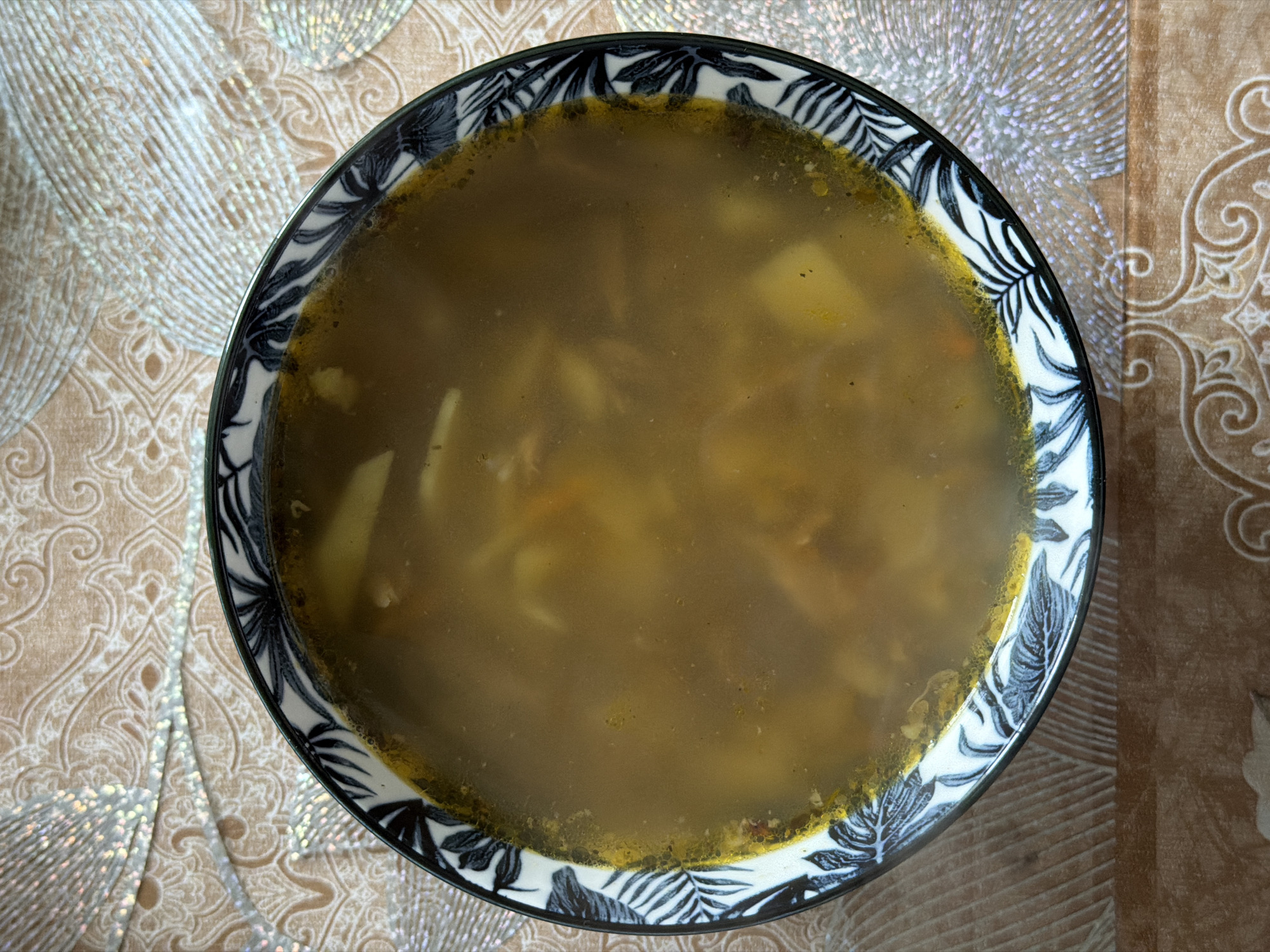 A bowl of soup sits on a patterned table in Заболотово, Russia. The soup is a murky brown color and appears to contain potatoes and possibly other vegetables. The bowl has a white, black, and blue leaf pattern. The table has a beige and brown patterned cloth with some silver accents.