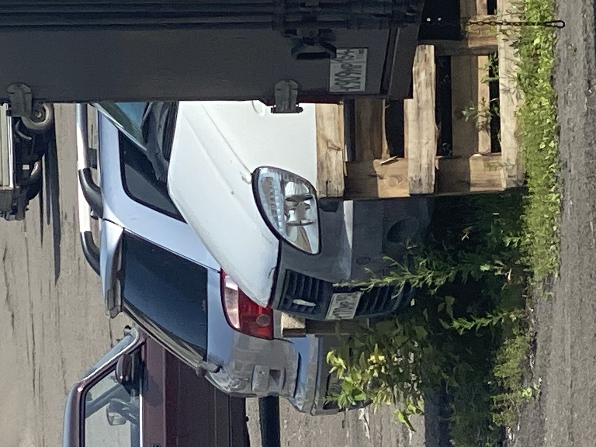 A close-up, eye-level shot captures the rear of a light-colored, possibly white, van or delivery vehicle partially obscured by a dark, ribbed vertical structure on the right side. The van's front right headlight assembly is visible, along with its grille and a license plate that appears to read "K909 MP 154". The vehicle is elevated by wooden pallets. To the left, a silver hatchback vehicle is visible, with its rear bumper and taillights in focus. Further left, the rear passenger side of a dark red sedan is partially seen. Green foliage, including small bushes and weeds, grows around the base of the vehicles and on the ground. The ground surface appears to be asphalt or a similar rough material. In the background, a light-colored truck with visible wheels is parked. The scene is likely an outdoor parking lot or industrial area. The license plate "K909 MP 154" is a Russian license plate, indicating the location is within Russia. Given the specific format of the license plate and the presence of other vehicles common in Russia, the scene is likely in Novokuznetsk, Russia.