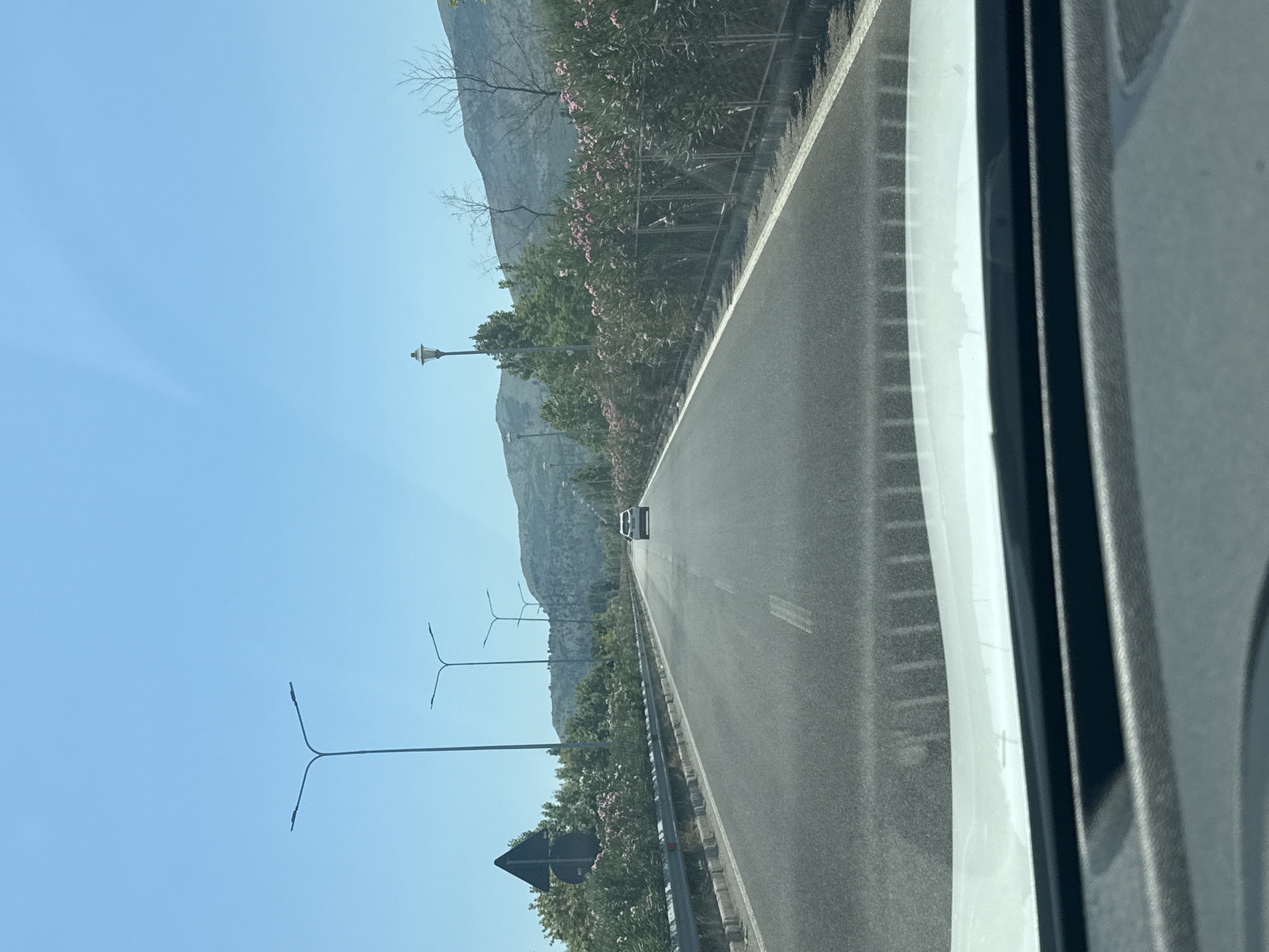The image depicts a perspective from inside a vehicle traveling on a road. The road itself is asphalt with white lane markings, and it extends into the distance. On the right side of the road, there is a metal fence bordering a line of dense green bushes interspersed with pink flowers. Beyond the bushes, a hill with sparse trees and dry branches is visible, leading up to a mountainous backdrop under a clear blue sky. On the left side of the road, there is a metal guardrail and a black triangular traffic sign. Several streetlights are visible along the roadside, with the closest ones on the left having a distinct Y-shaped design. Further down the road, a white car is seen driving ahead. The foreground shows the dashboard and windshield of the vehicle from which the photo is taken. The overall environment suggests a sunny day in a semi-rural or exurban setting, possibly on the outskirts of a city like Rrethinat, Albania, given the presence of road infrastructure and natural landscaping.
