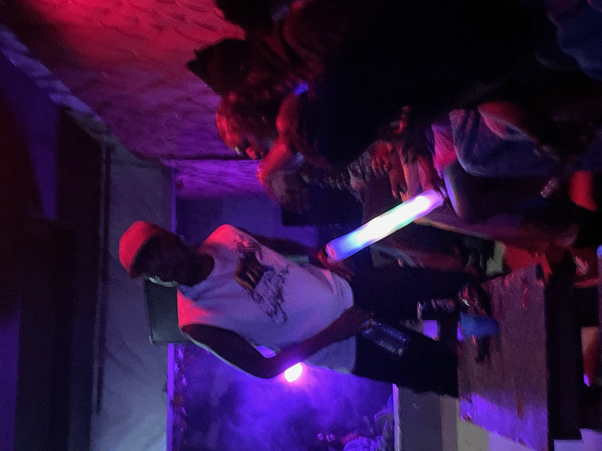 In Durumi, Nigeria, a dimly lit room, likely a nightclub or party venue, is illuminated by vibrant purple and red lighting, creating a hazy, atmospheric effect. In the foreground, a person wearing a red baseball cap and a white sleeveless shirt is holding a glowing, multi-colored light stick in their right hand and a bottle in their left. This individual appears to be interacting with others in the background, some of whom are obscured by shadows and the low lighting. A wooden table in the lower left corner of the image holds a blue cup and other indistinct objects. The overall scene suggests a lively social gathering.