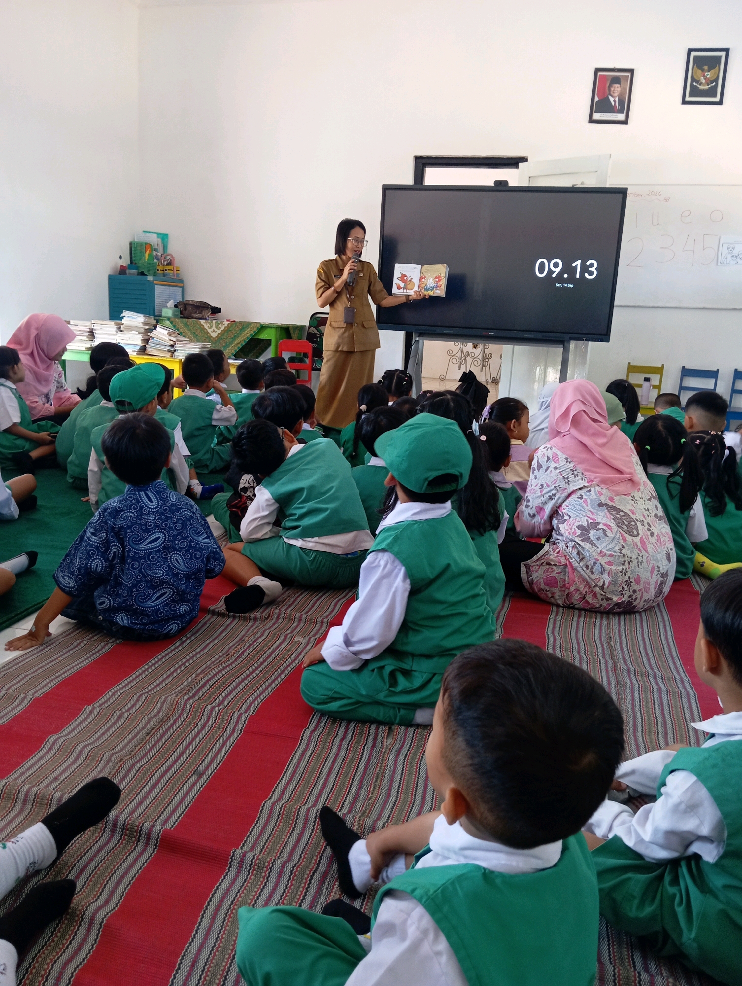 A teacher in a brown uniform is reading a book to a group of young children sitting on a striped mat in a classroom. The teacher is standing in front of a large interactive screen that displays the time as 09.13. The children are wearing green vests over white shirts and some have green caps. The classroom has white walls, a shelf with books and supplies, and framed pictures on the wall. Chairs are visible in the background. The overall impression is of an educational setting. Based on the style of the frames and the presence of Indonesian flags, it is likely located in Indonesia, possibly in Malang given the context.