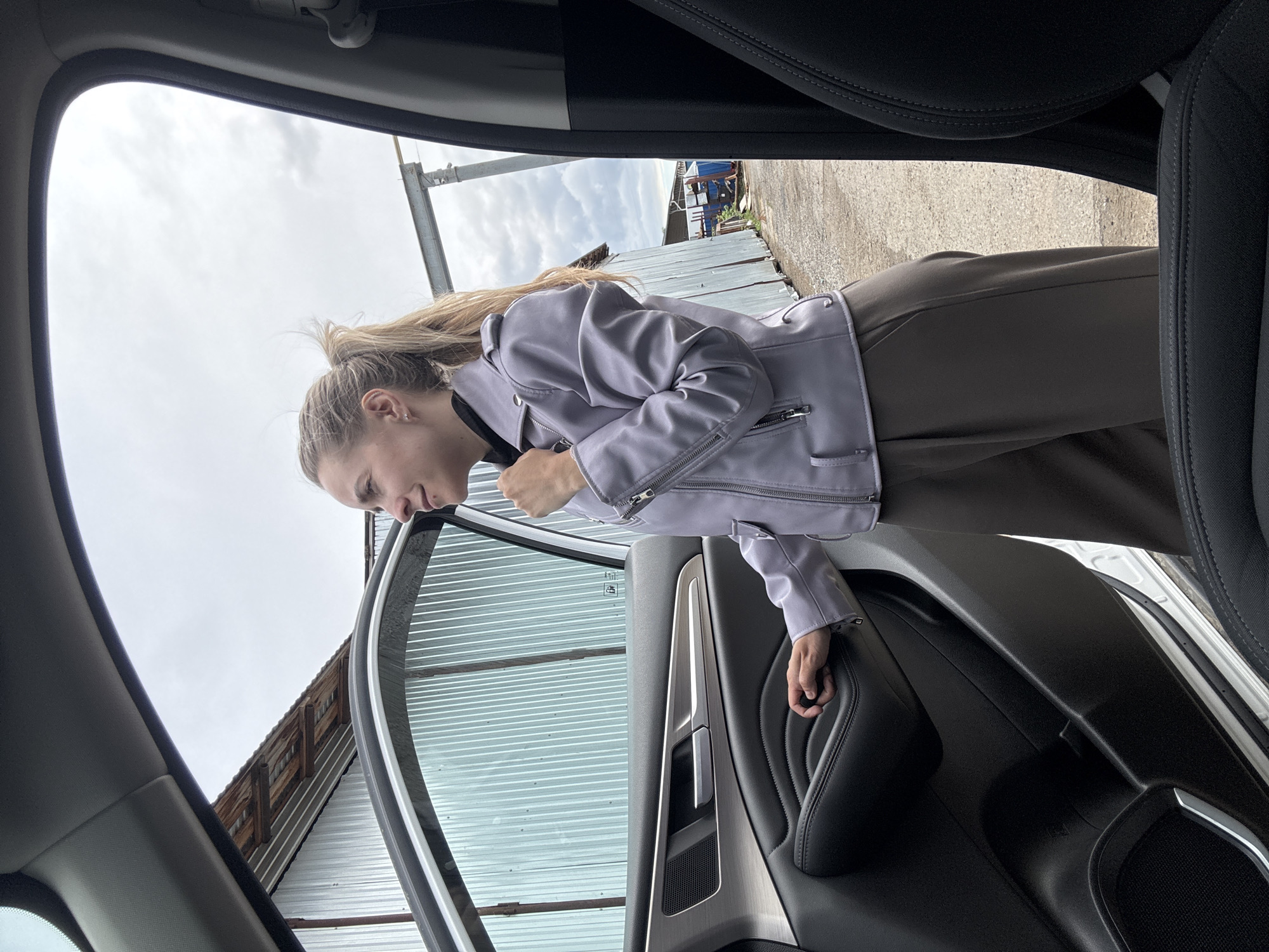 A person wearing a light purple jacket is positioned in the open doorway of a car. The person is leaning slightly into the doorway and looking downwards. Outside the car, there is a corrugated metal building and cloudy sky. The car's interior includes black leather seats and trim. The location is presumed to be near Chaikovsky, Russia based on user provided metadata.