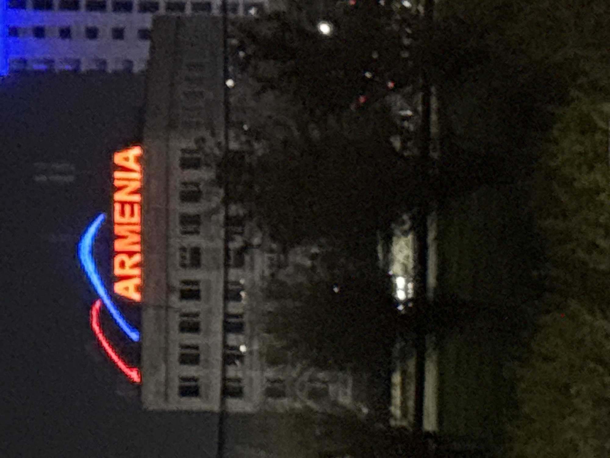 The image shows a nighttime view of a building in Yerevan, Armenia. The building has a sign on the top that says "ARMENIA" in orange neon letters, with blue and red neon arcs wrapping around the name. The building appears to be multi-storied, with multiple windows. To the right of the building is a dark area with what appears to be foliage and streetlights. Another building is visible to the left. The photo's angle suggests it was taken from below the building.