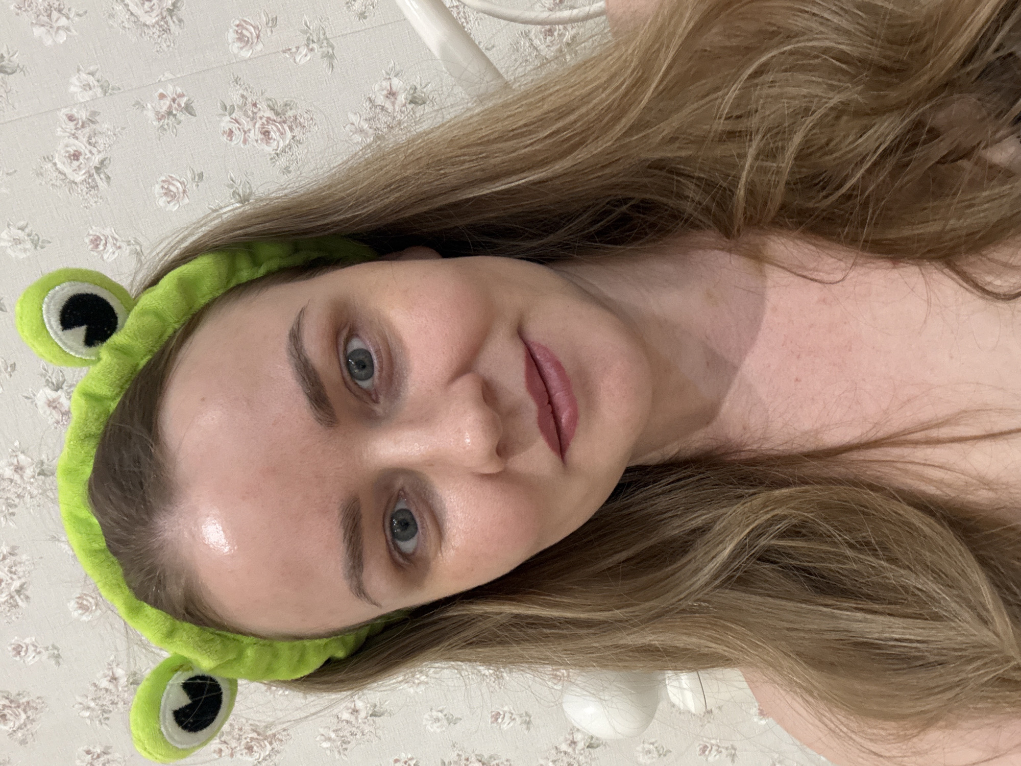 A close-up image shows a young woman with long, light brown hair, wearing a bright green, plush frog-themed headband. She looks directly at the camera with a calm expression and a faint smile. Her eyes are light-colored, and her lips are tinted with a soft pink or berry shade. Her skin appears somewhat dewy or hydrated on her forehead and nose. The background features light-colored wallpaper adorned with a subtle pattern of light pink and white roses. A white object, possibly furniture, is visible in the upper right background. The image is oriented sideways, implying the person is lying down or the photograph was taken from an unconventional angle. This scene takes place in Chaikovsky, Russia.