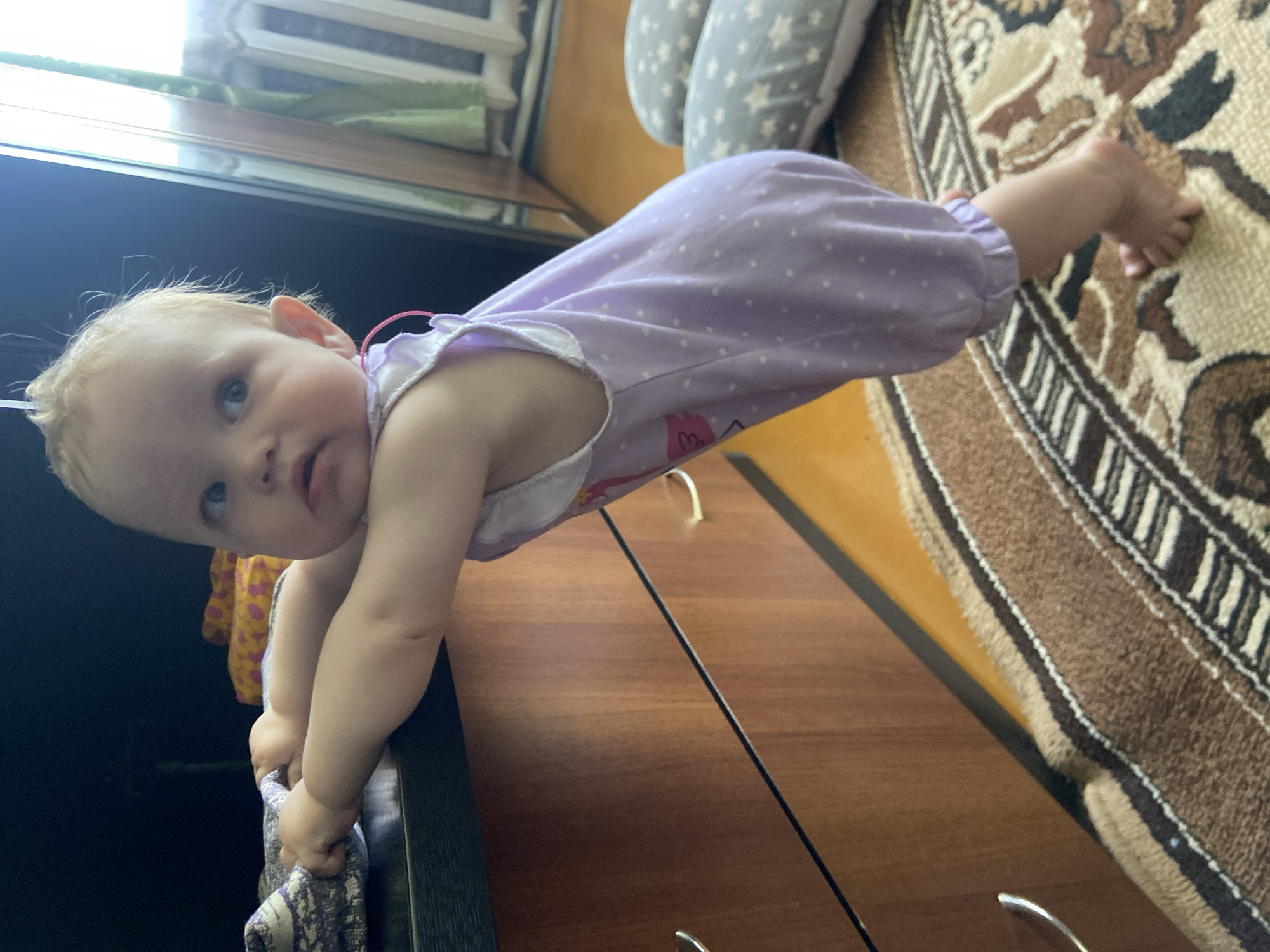 A child in a lavender polka-dotted romper is doing a plank on a brown wooden dresser in a room with a patterned rug. The child's feet are on the rug. The location is likely a home in Фоки, Russia.
