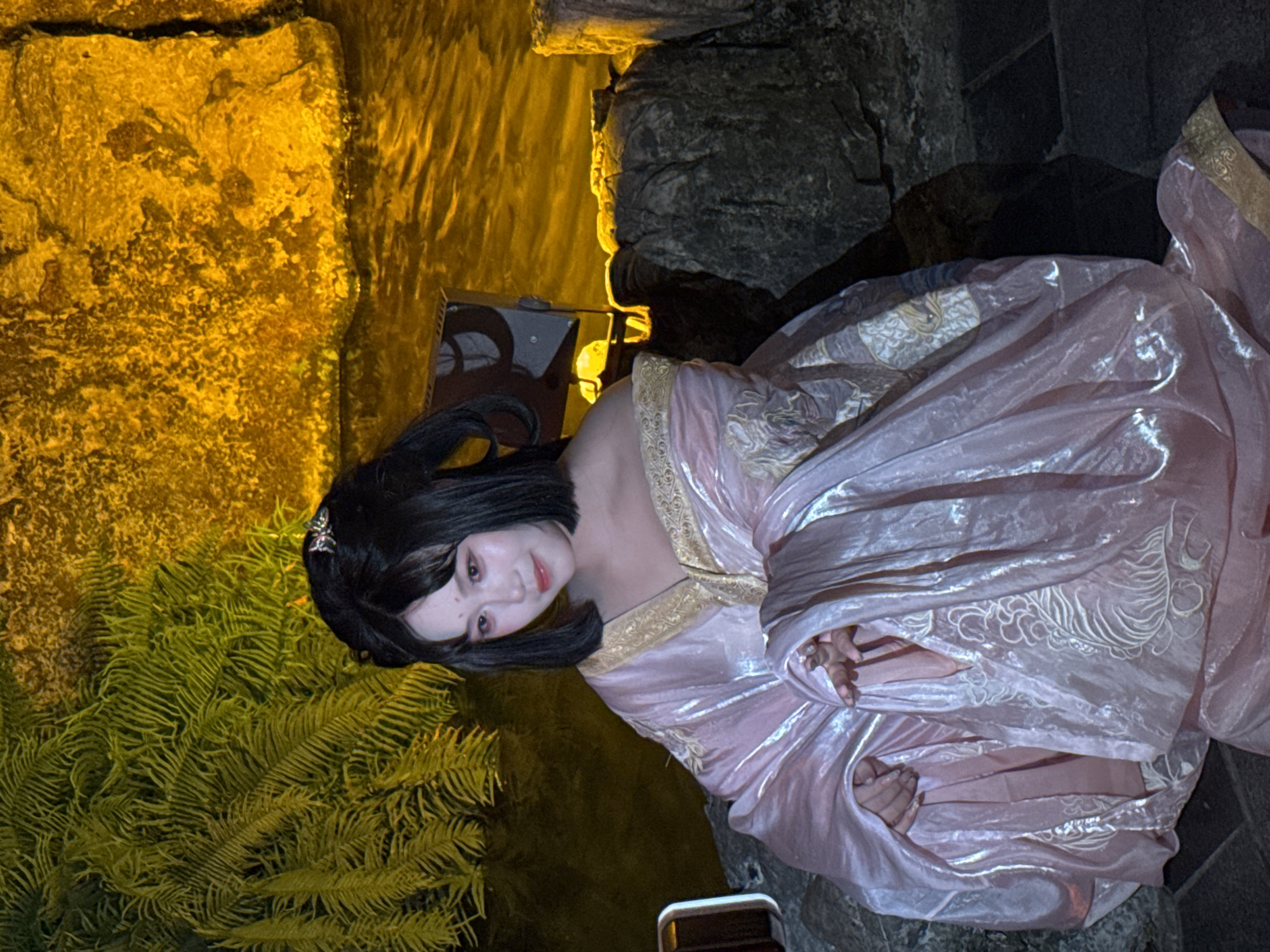 In Yuzhong District, China, a person adorned in a pink, traditional East Asian gown with golden embroidery is seated on rocks. The gown has flowing sleeves and intricate patterns of what appear to be mythical creatures or floral designs. The person has dark hair styled with a few ornaments and wears subtle makeup with red markings on their cheeks and forehead. Behind the individual, a body of water reflects a warm, yellow light, possibly from surrounding rocks or artificial lighting, creating a tranquil yet dramatic ambiance. Lush green foliage, likely ferns, is visible to the left, and more rocks are scattered around the scene. The overall impression is one of serene beauty and cultural richness, possibly a staged performance or a themed photoshoot set in a garden or near a water feature.