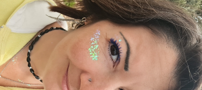 The image presents a close-up, right-side portrait of an individual, taken outdoors in the Mate Asher Regional Council, Israel. The subject, appearing female, displays dark brown hair, a brown eye with vibrant blue mascara on the eyelashes, and a defined dark eyebrow. Iridescent glitter is applied to the upper right cheekbone, forming two distinct clusters: one pinkish-white near the temple and another light green below the visible eye. A silver stud pierces the right nostril. The right earlobe is adorned with an earring featuring multiple dangling silver chains. The individual wears a yellow hooded garment with a visible white collar. A black beaded choker necklace, interspersed with small silver-colored spacers, is worn around the neck, along with a thinner silver chain necklace that has a partially visible pendant. The background consists of blurred green foliage, consistent with an outdoor environment.