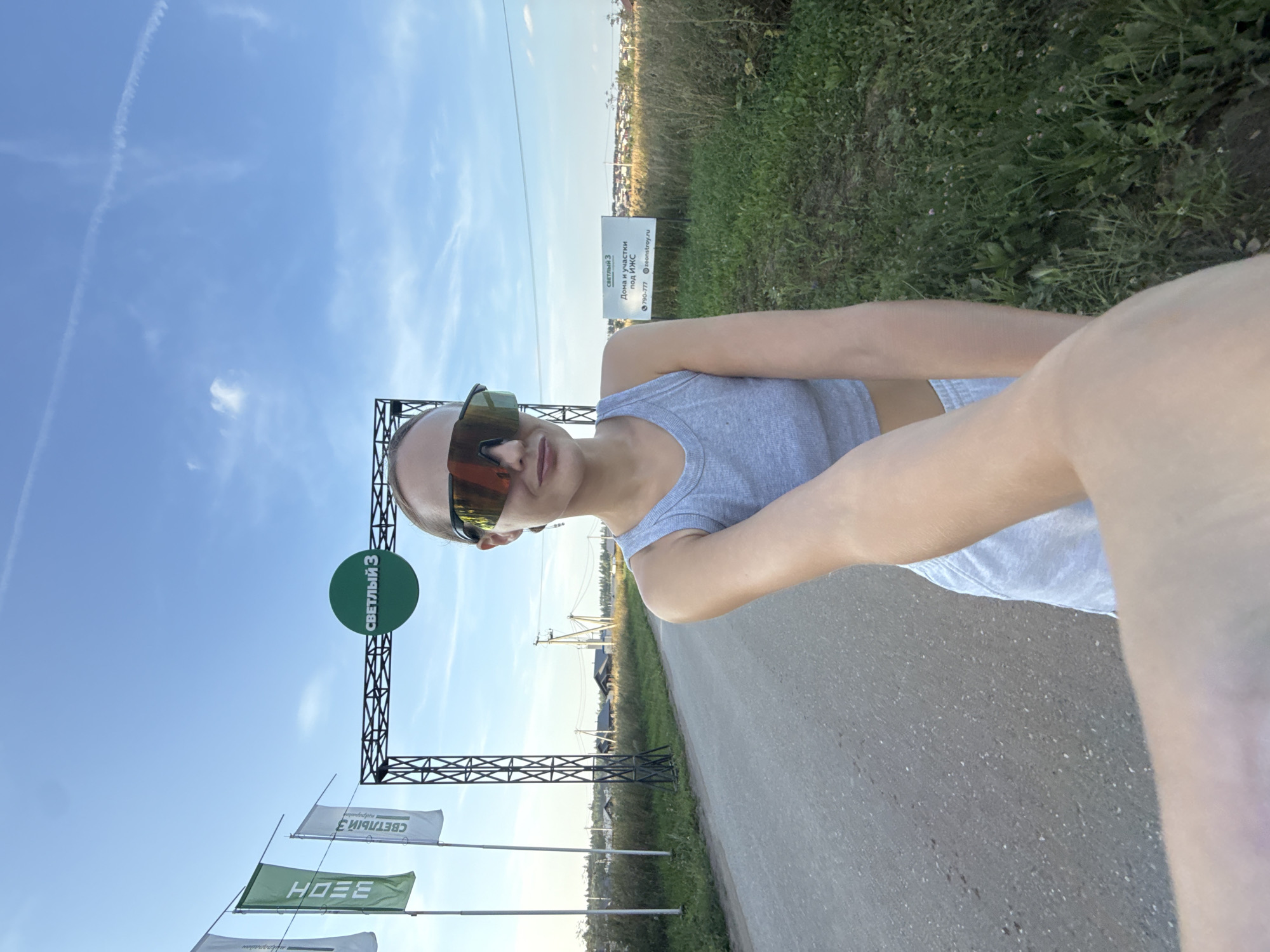 A woman wearing a grey tank top and grey shorts, along with large, mirrored sunglasses, stands on a dirt road. She is holding a camera, taking a selfie. In the background, a black metal archway with a green circular sign reading "СВЕТЛЫЙ 3" is visible. To the left of the archway are three tall flags. The flag on the far left is white with green lettering. The middle flag is green with white lettering that appears to read "ЗЕОН". The flag on the far right is white with green lettering. Behind the flags and archway, there is a grassy field with scattered houses and utility poles. To the right of the archway, a white sign with black text reads "СВЕТЛЫЙ 3 ДОМА И УЧАСТКИ ПОД ИЖС" and a phone number and website address. The sky above is blue with some white clouds and a faint contrail from an airplane. The location is identified as the village of Podshivalovo, Russia.
