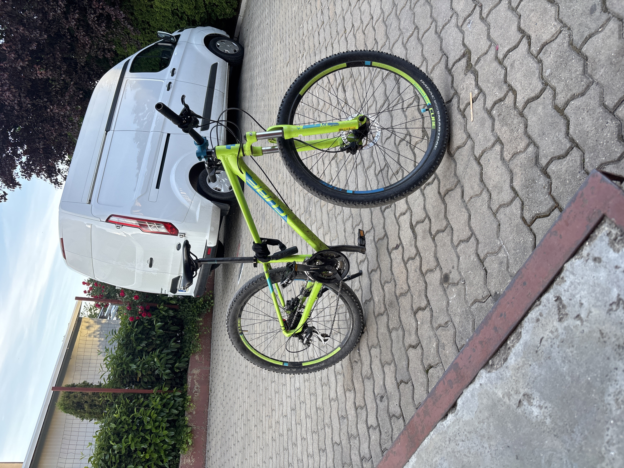 A lime green and blue CUBE mountain bike stands on a gray brick pavement next to a white van. Behind the van, a bush with red roses is visible near a metal fence. The location is in the city of Unknown, Unknown.