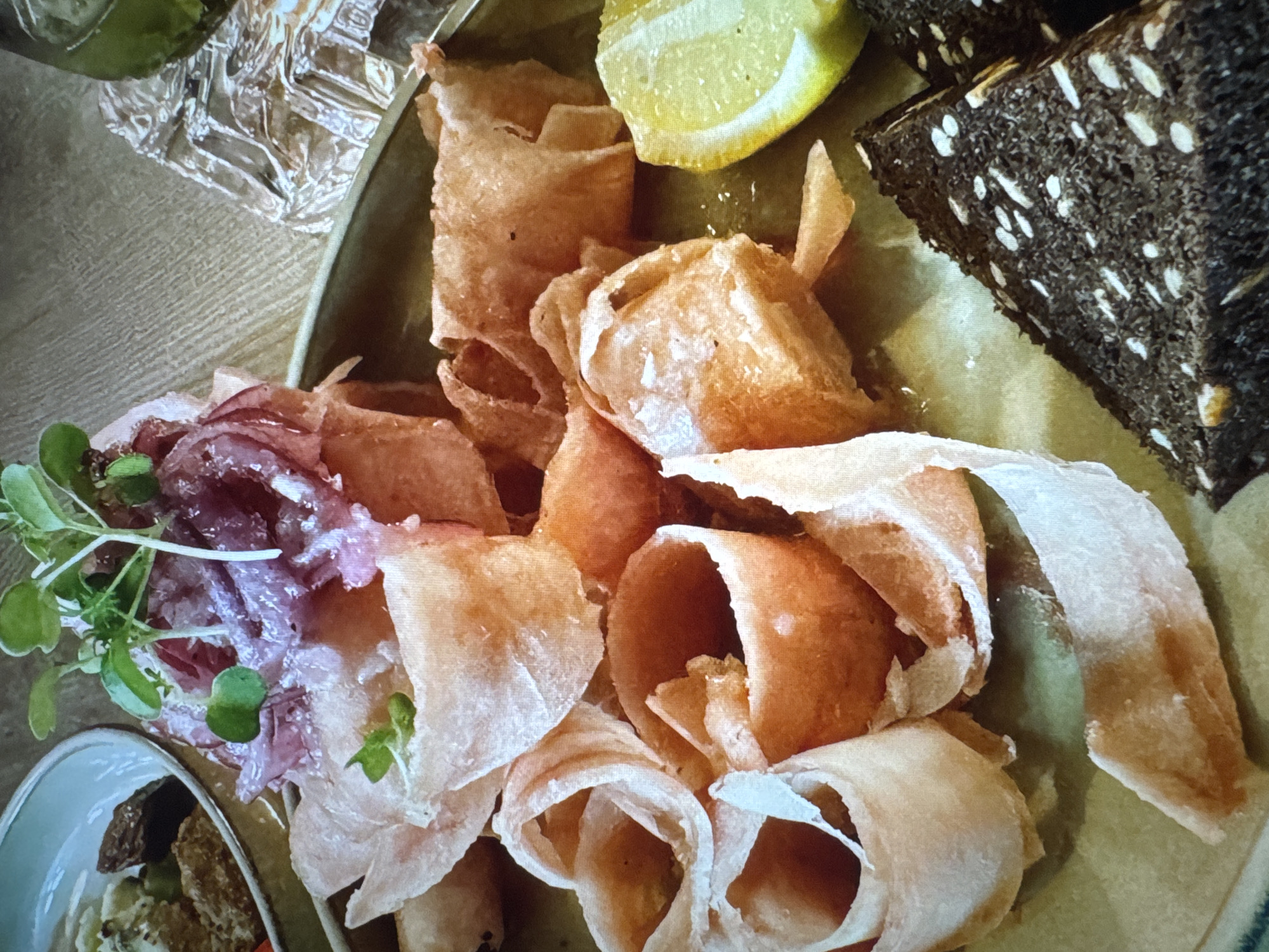 A close-up, eye-level shot shows a plate of food. The plate appears to be a pale green ceramic dish. Dominating the center of the plate are several pieces of thinly sliced, pinkish-orange food, curled into tube-like shapes, some appearing slightly fried or crispy. On top of these curled pieces, a small mound of thinly sliced red onion is visible, garnished with fresh green sprouts. To the right of the curled food is a wedge of bright yellow lemon. In the bottom right corner, two triangular slices of dark brown bread, studded with white seeds, are arranged. In the background, a portion of a white bowl containing a light-colored food item and some dark pieces can be seen. The surface beneath the plate is a light brown, textured material, possibly wood.