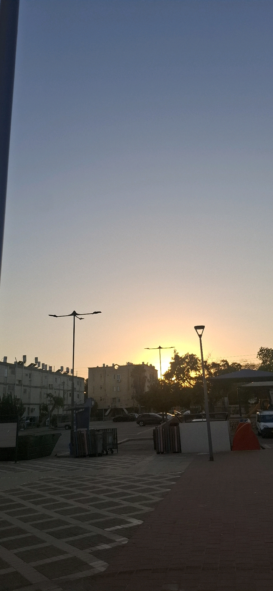 The image depicts a street scene in Yerocham, Israel, during sunset. The sky is a gradient of pale blue and soft orange, indicating the setting sun. In the foreground, a patterned paved area is visible, transitioning into a darker paved surface. To the left, there are large green bins, and a blue recycling bin with metal carts. Further back, several apartment buildings with flat roofs and visible air conditioners are lined up. Across the street, more buildings of varying heights and architectural styles are present, with trees and foliage interspersed. Two streetlights with multiple lamps are prominent in the mid-ground, silhouetted against the bright sky. Cars are parked in a lot behind the buildings. A modern, light-colored lamp post stands on the right side of the image, and a bright orange structure, possibly a safety barrier or a piece of equipment, is situated near a white wall. The overall atmosphere is one of a typical urban residential area at dusk.