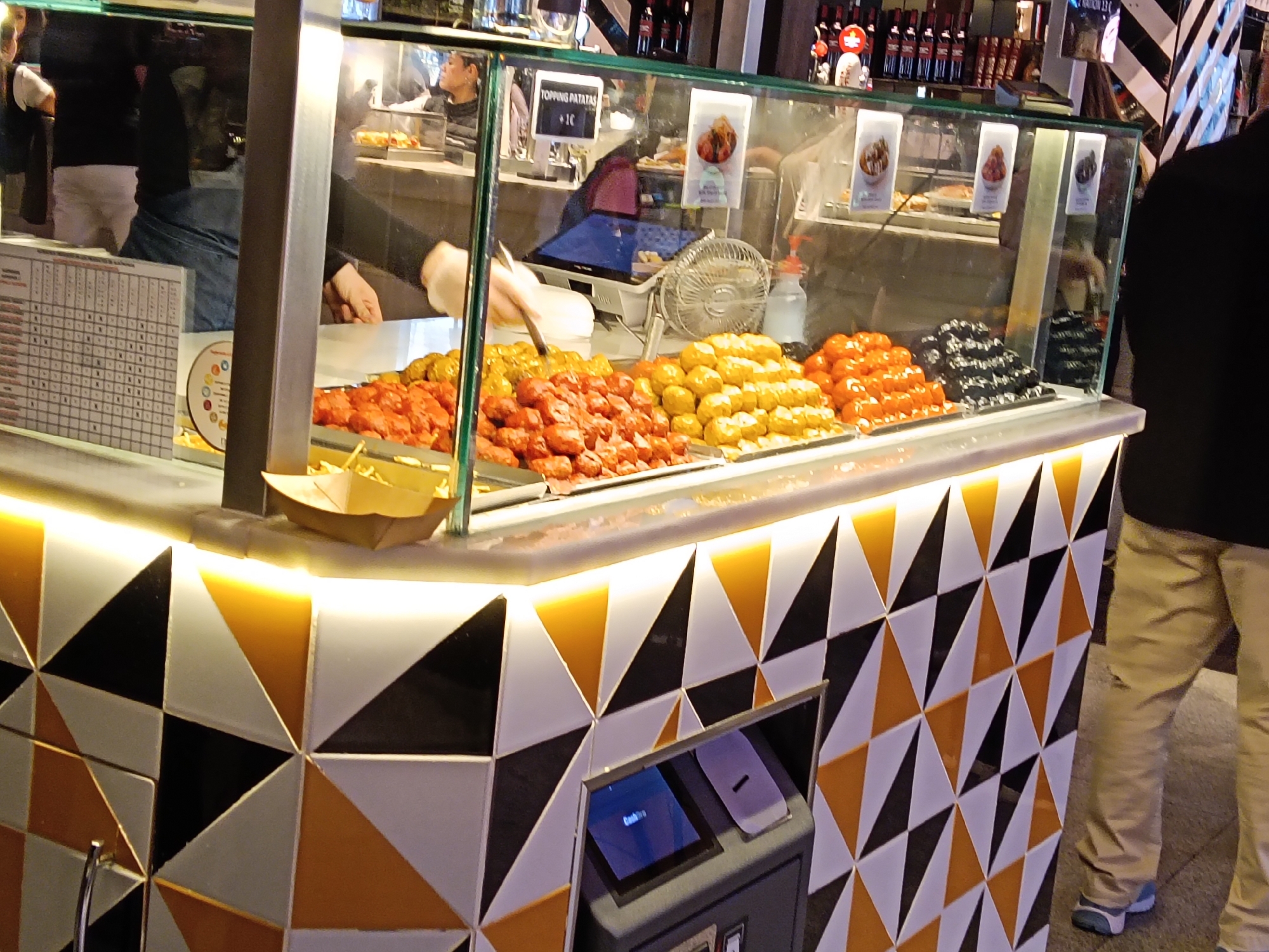 The image depicts a food stall in Madrid, Spain, likely within a market or food hall. The stall is enclosed in a glass display case, showcasing a variety of "patatas bravas", a common Spanish tapa of fried potatoes served with different sauces. Four distinct variations of patatas bravas are visible.
Behind the food items, an employee wearing gloves is seen preparing or serving the food. A cash register and a small fan are also located on the counter behind the food displays. On the display case, there is a sign indicating that topping patatas costs 1 Euro.
The exterior of the stall features a geometric pattern of alternating black, white, and yellow triangles. A payment terminal for card transactions is built into the base of the stall. A person wearing khakis is standing in the corner of the frame, implying the scene is a casual and bustling eatery.