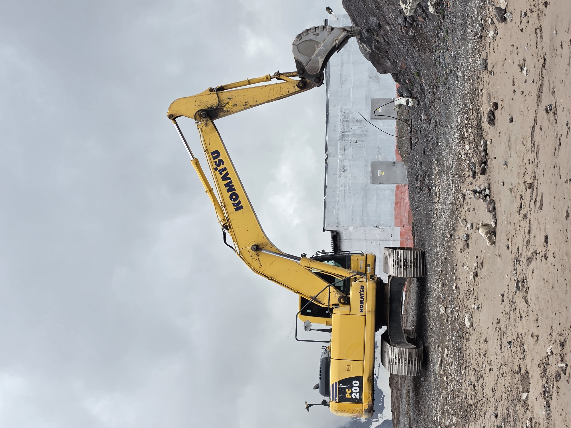 An excavator, identified by the "KOMATSU" logo and "PC 200" model number, is positioned on a muddy, rocky terrain. The excavator is yellow with a black cabin and is equipped with a large bucket. It is actively engaged in moving a pile of dark, rocky material. In the background, a light-colored building with two doors is visible. The sky is overcast with clouds. The scene suggests a construction or excavation site. The location is identified as Terskol, Russia, near a mountainous region, as indicated by snow-capped mountains in the far left background.