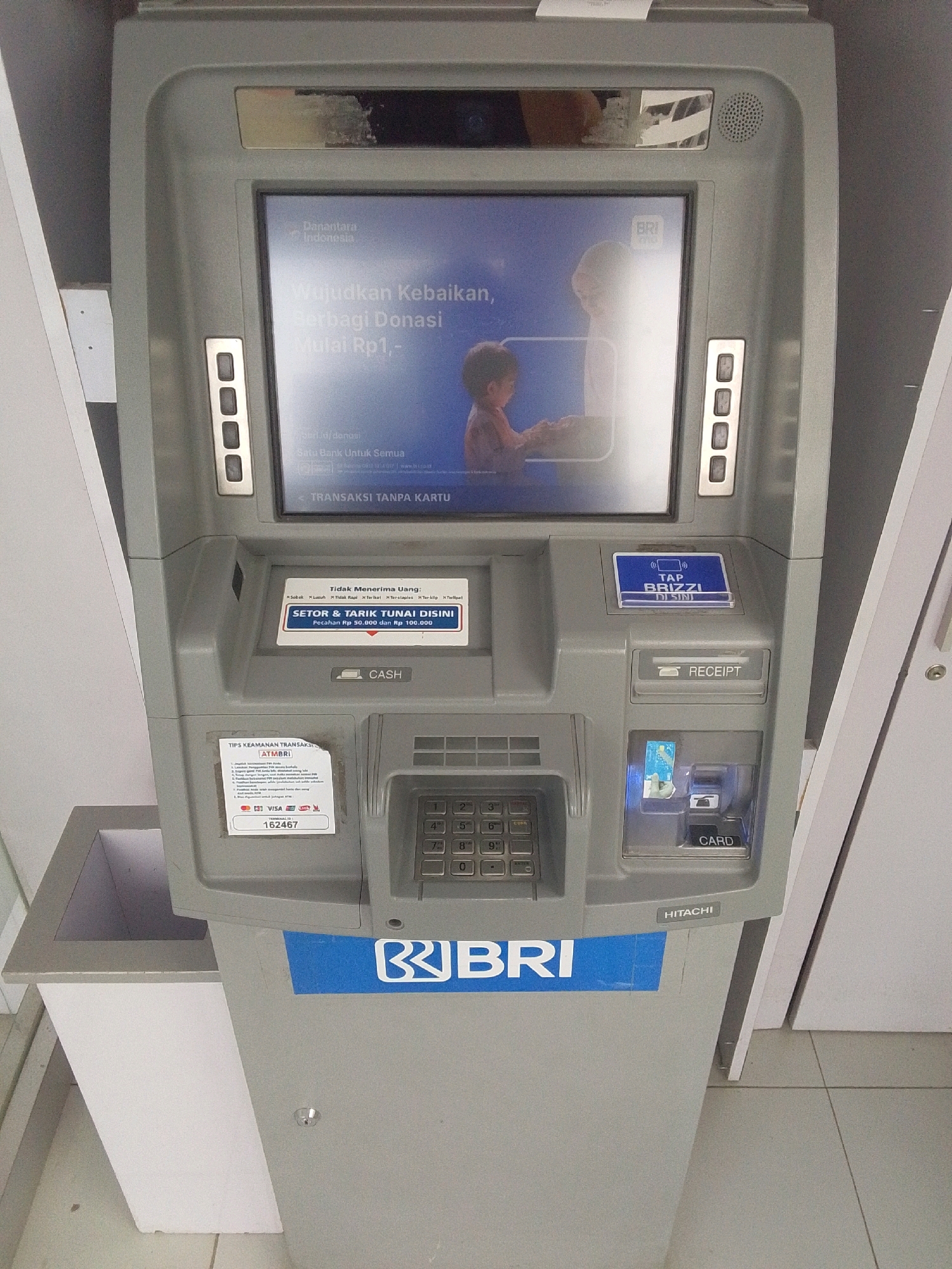 This is a close-up, eye-level, front-facing shot of a BRI (Bank Rakyat Indonesia) ATM machine located in Purworejo, Indonesia. The ATM is gray with a screen displaying a blue background with text in Indonesian about donations. There are several buttons on the left and right sides of the screen. Below the screen, there is a slot for cash deposits and withdrawals, a receipt dispenser, and a card slot. A sticker with security tips for ATM transactions is visible on the lower left. A keypad is located to the right of the cash slot. The ATM is branded with the BRI logo. The background is blurred, but appears to be an indoor setting with a tiled floor and white walls.
