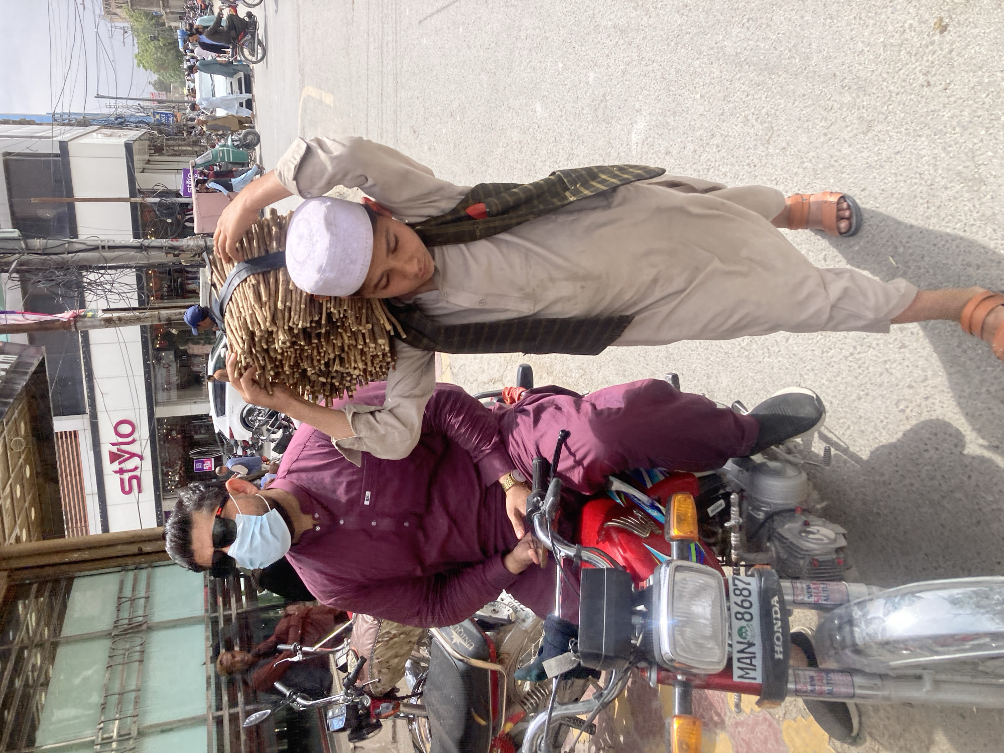 The image shows two men on a motorcycle on a street in Quetta City Tehsil, Pakistan. One man is driving the motorcycle, wearing a dark purple kurta and a face mask. The other man is lying across the seat of the motorcycle, carrying a bundle of sticks on his head. He is wearing a light-colored robe and a white cap. The motorcycle has a license plate that reads "MAN 8687". In the background, there are buildings and other people on the street. One of the buildings has a "stylo" sign on it. Overhead are many electrical wires.