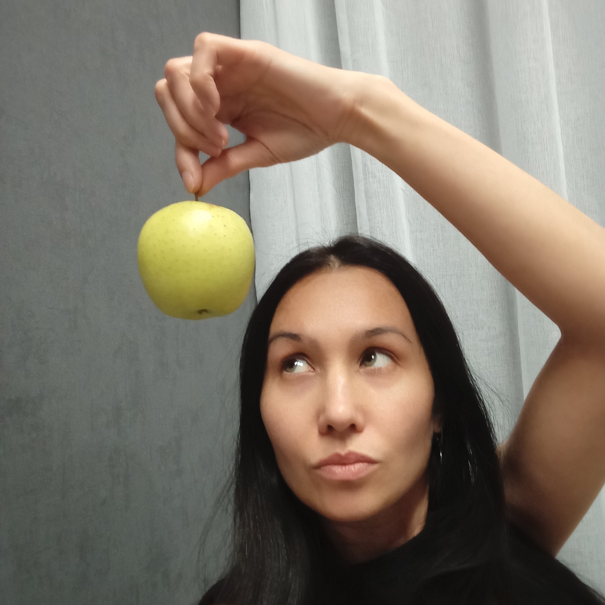 A woman with dark hair holds a green apple in her right hand above her head. She is looking up at the apple with a thoughtful expression. The background consists of a gray wall on the left and a white curtain on the right.
There is no information available about the location of the image being in Pervomaysky, Russia.