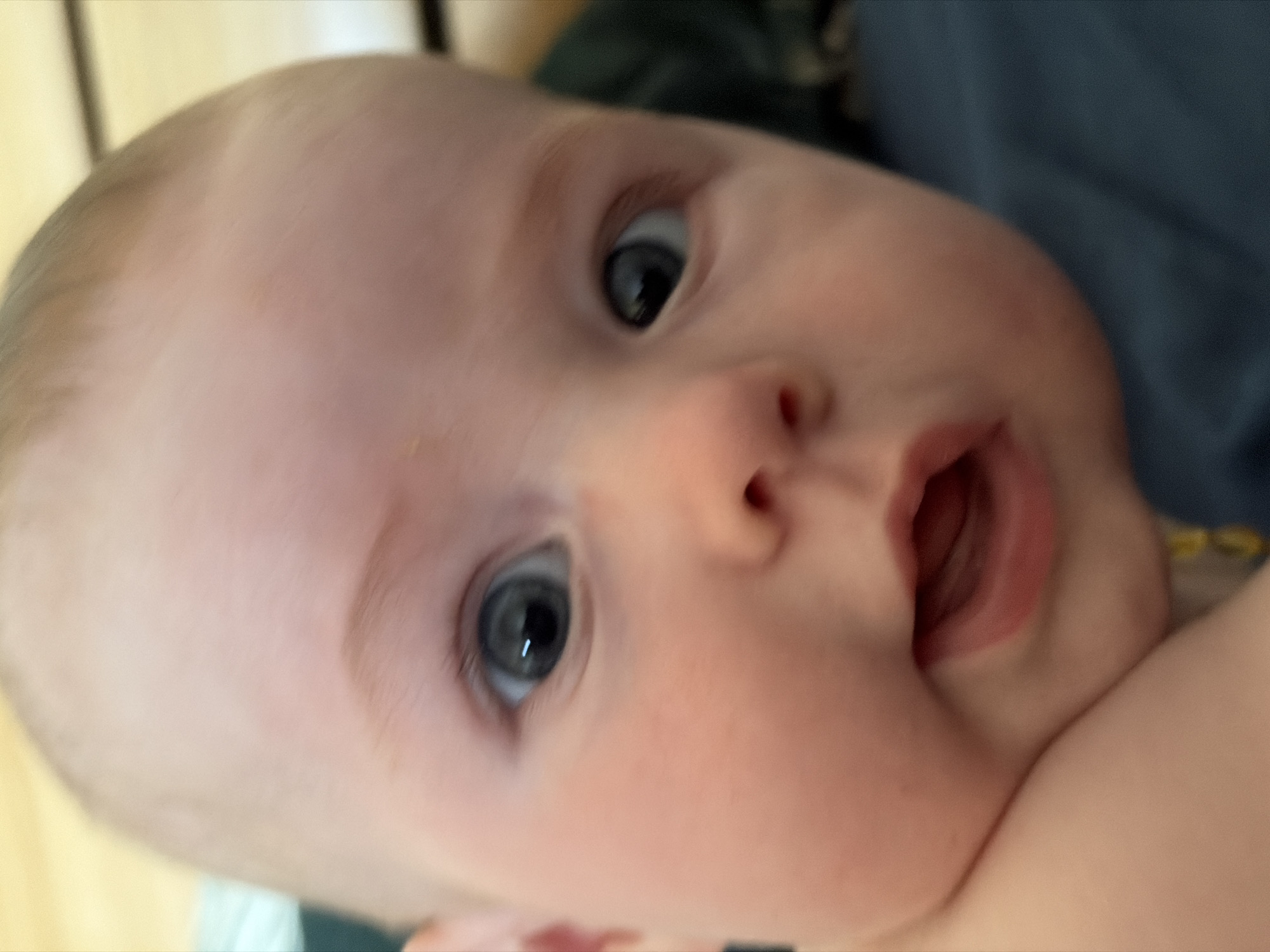 Close-up, blurred image of a baby with fair skin, greyish-blue eyes, and sparse light hair. The baby's mouth is slightly open. The background is out of focus, revealing a light-colored wall on the left and a dark blue garment on the upper right. The presence or absence of context connecting this image to Chaikovsky, Russia is unknown.
