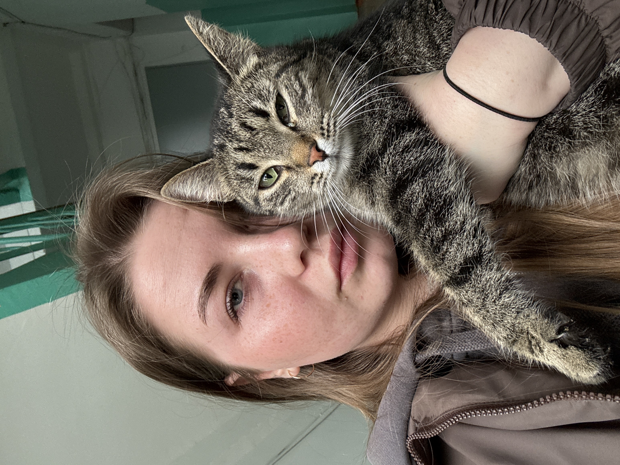 A young woman with light brown hair and blue eyes is holding a tabby cat against her face. She has fair skin with some freckles, and is wearing a brown jacket and a black choker necklace. The cat has green eyes and prominent white whiskers. The background is out of focus but appears to be an interior space with a white door or wall. There is no clear indication of the location being Chaikovsky, Russia.