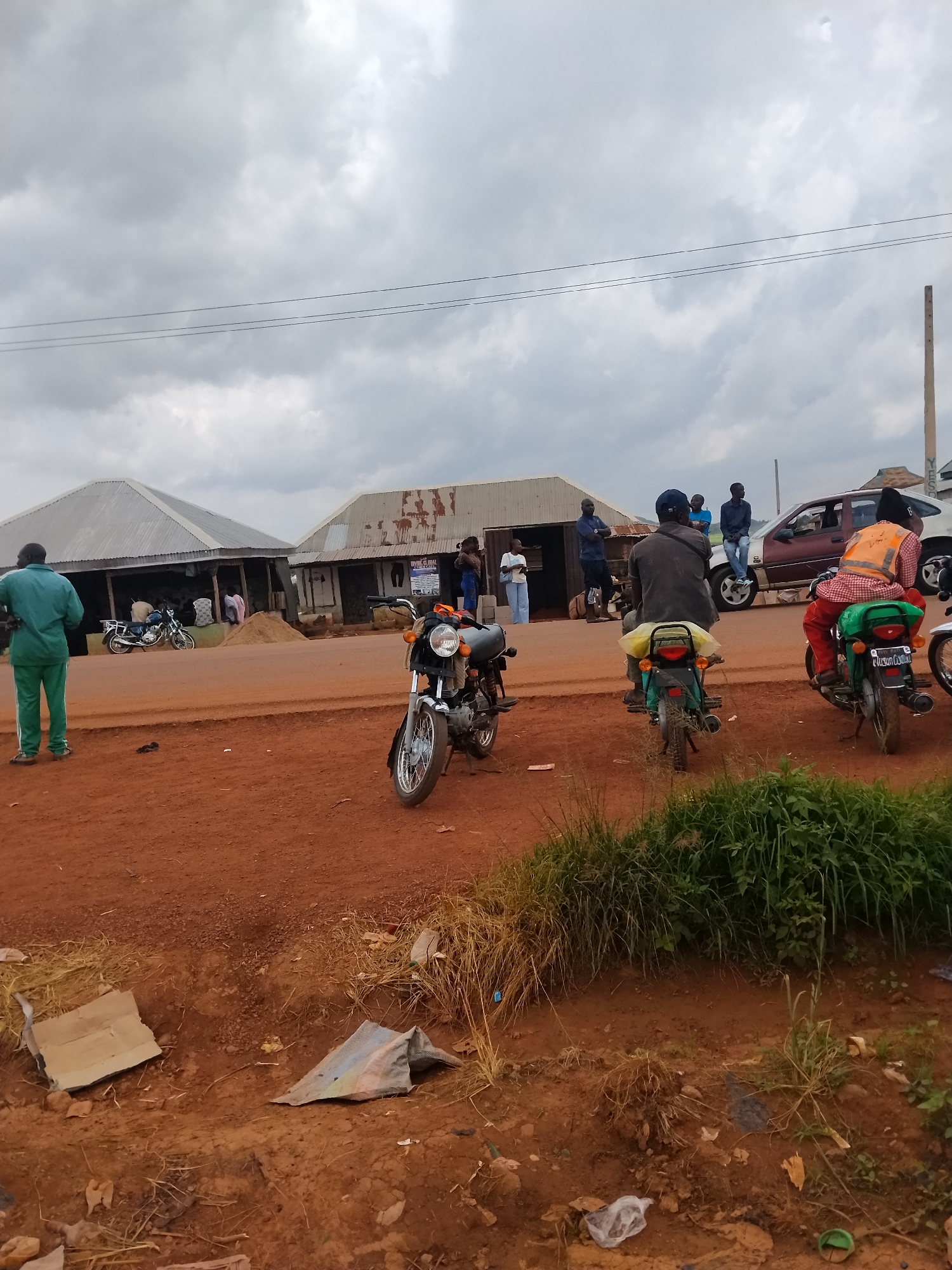 The image captures a street scene in Durumi, Nigeria, with a focus on a group of people and motorcycles. The environment is characterized by a dirt road, a cloudy sky, and modest roadside structures that appear to be shops or market stalls with corrugated metal roofs.
In the foreground, a motorcycle is parked on the right, facing left. Further back, a man in a green tracksuit stands on the left, facing away from the camera. Several other motorcycles are present, some with riders. One motorcycle on the left has a rider in a black jacket, and another motorcycle in the center has a rider wearing a black cap and a yellow bag on the back. To the right, a motorcycle with a rider in an orange reflective vest and a red and white checkered shirt is also visible.
A car, predominantly maroon with a white roof, is parked behind some of the motorcycles. Several individuals are standing around the shops, with some appearing to be engaged in conversation or observing the surroundings. One person is standing near a pile of sand.
The atmosphere appears to be overcast, contributing to a subdued lighting condition. The ground is primarily composed of reddish-brown dirt, with patches of dry grass and some scattered debris, including what looks like discarded cardboard and plastic. The buildings in the background are simple, with open fronts suggesting a market or informal business area. The overall scene depicts a typical day in a Nigerian community, with transportation and commerce being central elements.