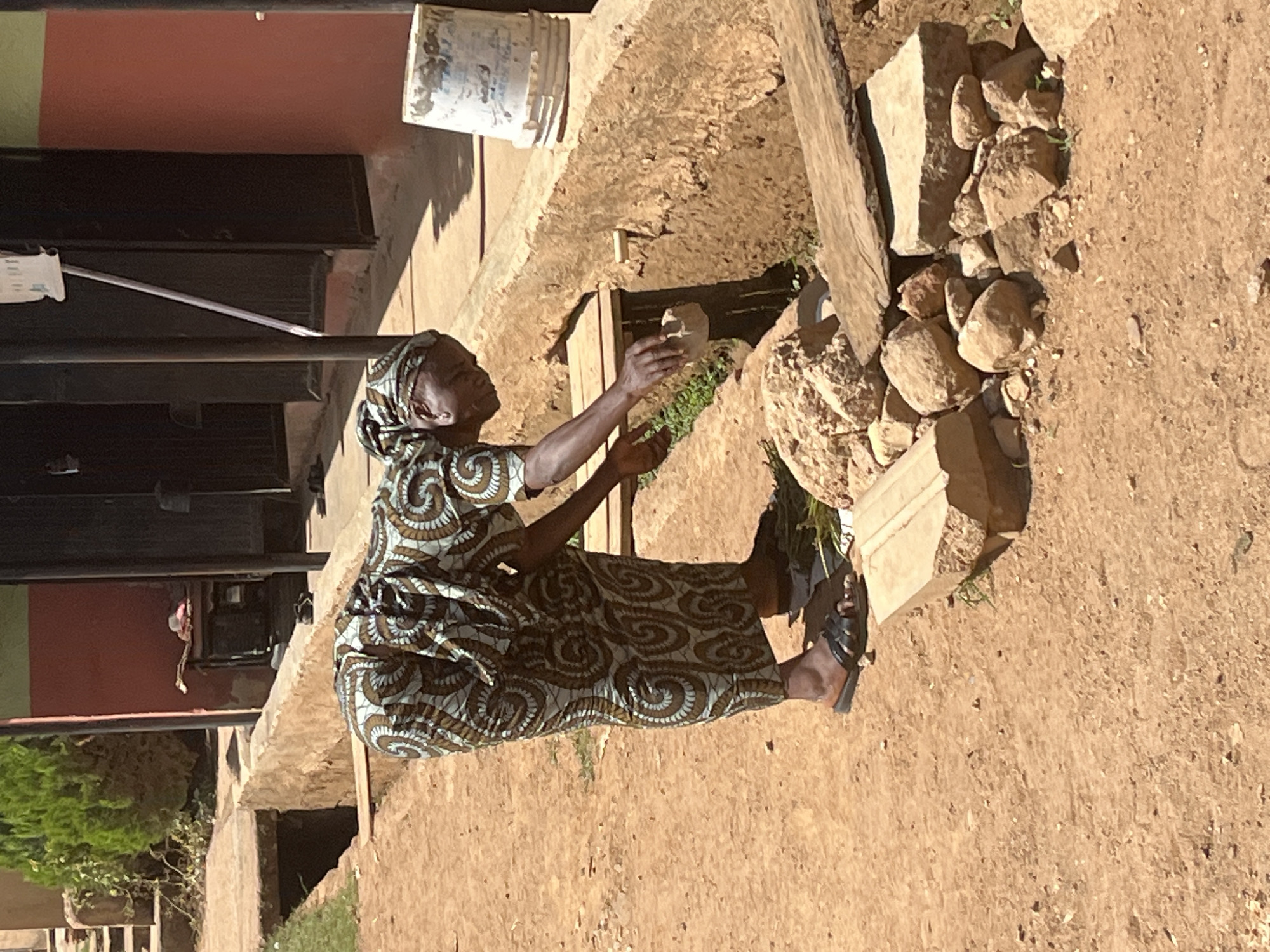 An older woman in a patterned dress and headscarf is squatting outdoors, holding a rock in her outstretched hand. She is surrounded by other rocks and stone building materials. The setting appears to be a construction or renovation site, with parts of a building visible in the background. The location is Barakin Akawo, Nigeria.