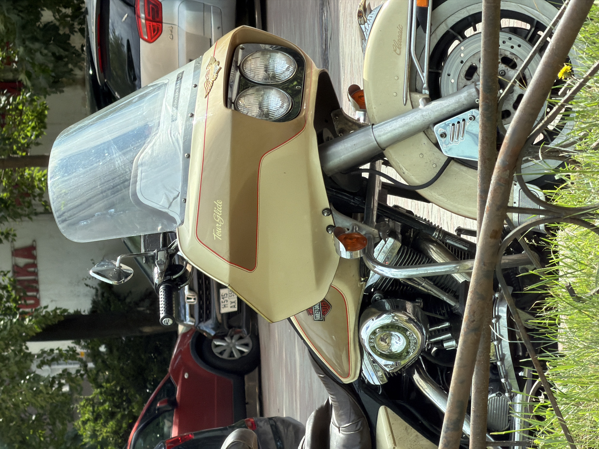 This is a close-up, eye-level, outdoor, medium shot of a beige Harley-Davidson motorcycle parked on a sidewalk, as indicated by the presence of grass and a rusty metal fence in the foreground. The motorcycle is the primary subject, facing towards the viewer's left. Its front fairing, windshield, dual headlights, and part of the engine are visible. The fairing is cream-colored with a brown stripe and red pinstriping, featuring the "Tour Glide" text and an eagle emblem. A Harley-Davidson logo is visible on the side of the fairing. The motorcycle's engine components, chrome exhaust pipes, and a portion of the seat are also in view. In the background, there are parked cars, including a red car and a silver car, and foliage. A building with red Cyrillic lettering, which reads "ПОДРУЖКА," is visible in the upper left quadrant, indicating the location is Saint Petersburg, Russia. The lighting suggests it is daytime, with sunlight illuminating the scene. There are no people visible in the frame.
