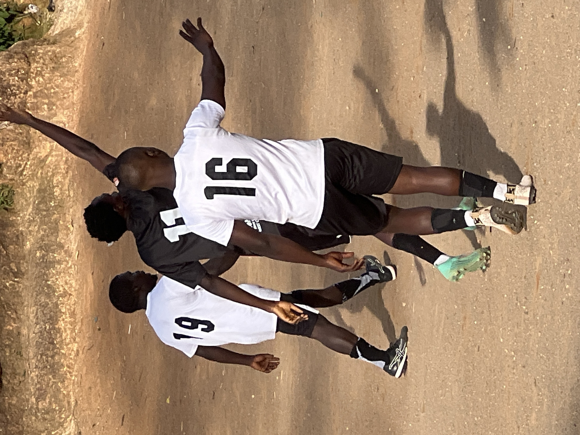 The image depicts a scene outdoors, likely in Jos, Nigeria, with a sandy or dirt ground. Three individuals, appearing to be young men, are engaged in a sporting activity, possibly a soccer game. They are wearing athletic attire including jerseys and shorts.
The person in the foreground, facing away from the camera, is wearing a white jersey with the number "16" in black on the back. They are also wearing black shorts and black socks with white and grey accents, along with athletic shoes. This individual's right arm is extended outwards to the right.
To the left of the foreground person, another individual is wearing a white jersey with the number "19" in black on the back, black shorts, and black socks with grey and white accents. Their left arm is partially visible.
In the background, a third individual is wearing a dark jersey, possibly black, and appears to be reaching upwards with their right arm.
The ground is a mix of sand and dry vegetation. The lighting suggests it is daytime, with shadows cast on the ground indicating the presence of the sun. The background shows a rocky or embankment area with some sparse vegetation. The overall impression is an informal sports setting in a relatively dry, open environment.