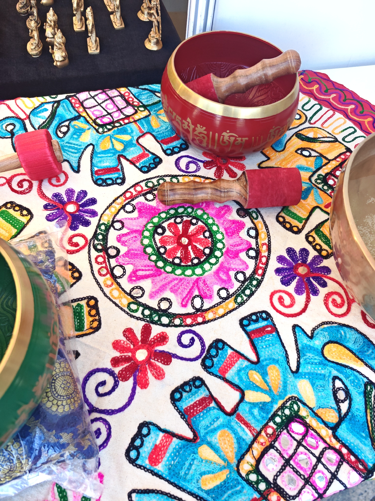 The image depicts a close-up view of a table adorned with colorful, embroidered fabric. The fabric features intricate patterns, including elephants, flowers, and geometric designs, with small mirrors incorporated into some of the motifs. Several singing bowls and their associated mallets are placed on the fabric. One red singing bowl with golden inscriptions is prominent, containing a wooden mallet with a red fabric tip. Another mallet with a similar red tip rests on the fabric. A green singing bowl is partially visible on the left side of the frame, and a portion of a brass singing bowl is seen on the right. In the background, on a dark surface, are several small, golden figurines, possibly decorative statues or chess pieces. The lighting suggests an indoor setting, likely a market stall or shop. There are no people visible in the image, and the scene appears to be static. The visual style is bright and detailed, emphasizing the textures and colors of the embroidered textile and the metallic sheen of the bowls and figurines. There is no information in the image to suggest a specific location in Sofia, Bulgaria.