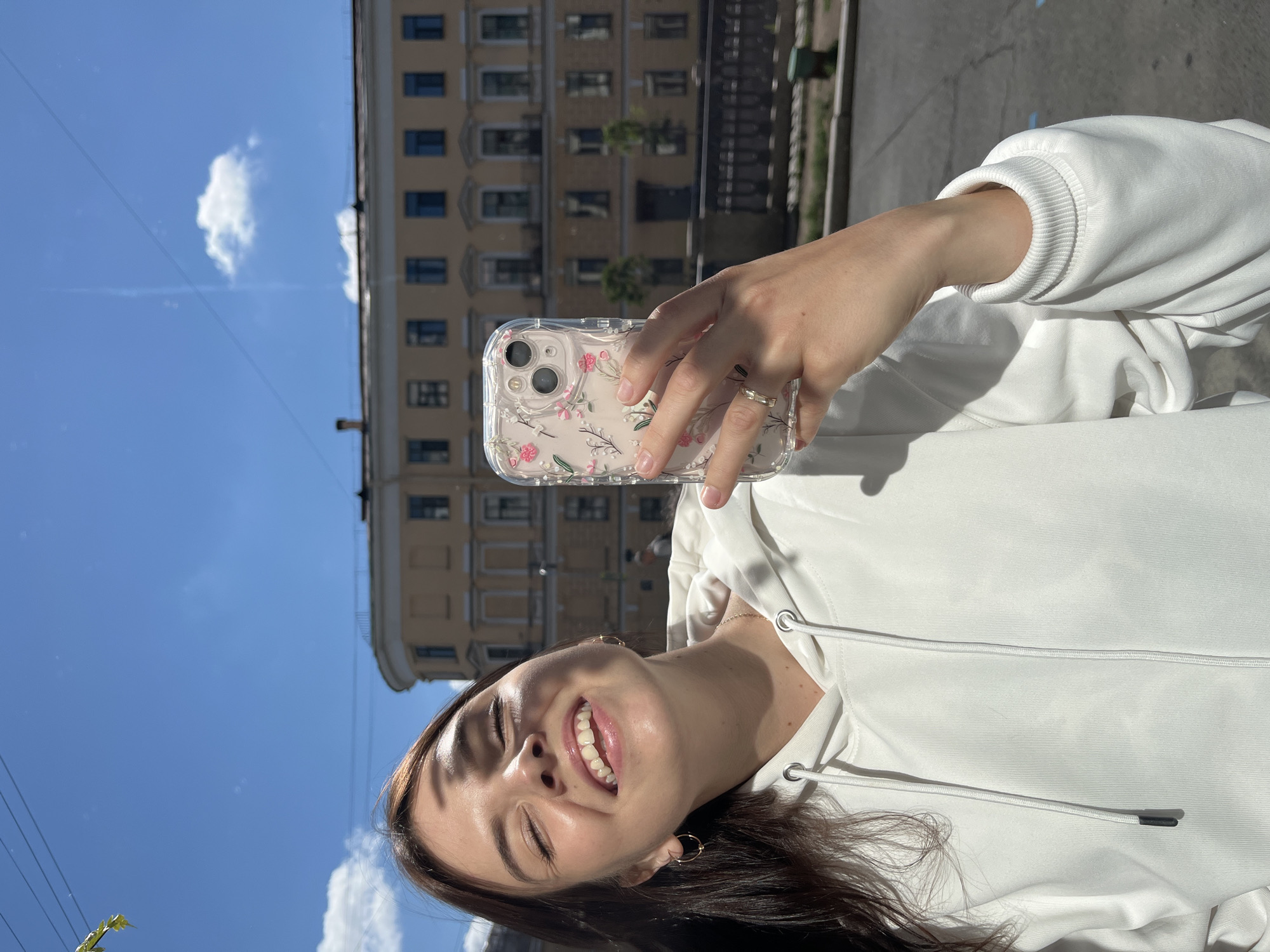 A young woman with dark hair is taking a selfie outdoors on a sunny day in Saint Petersburg, Russia. She is wearing a white hoodie and gold hoop earrings. She is smiling with her eyes closed, her face illuminated by the sunlight casting a shadow across her forehead. In her right hand, she holds a clear phone case with a floral pattern. Behind her, a multi-story building with yellow facade and numerous windows is visible, characteristic of Saint Petersburg's architecture. The sky is blue with scattered white clouds. The street in front of the building appears to be paved, with a low railing along a sidewalk.