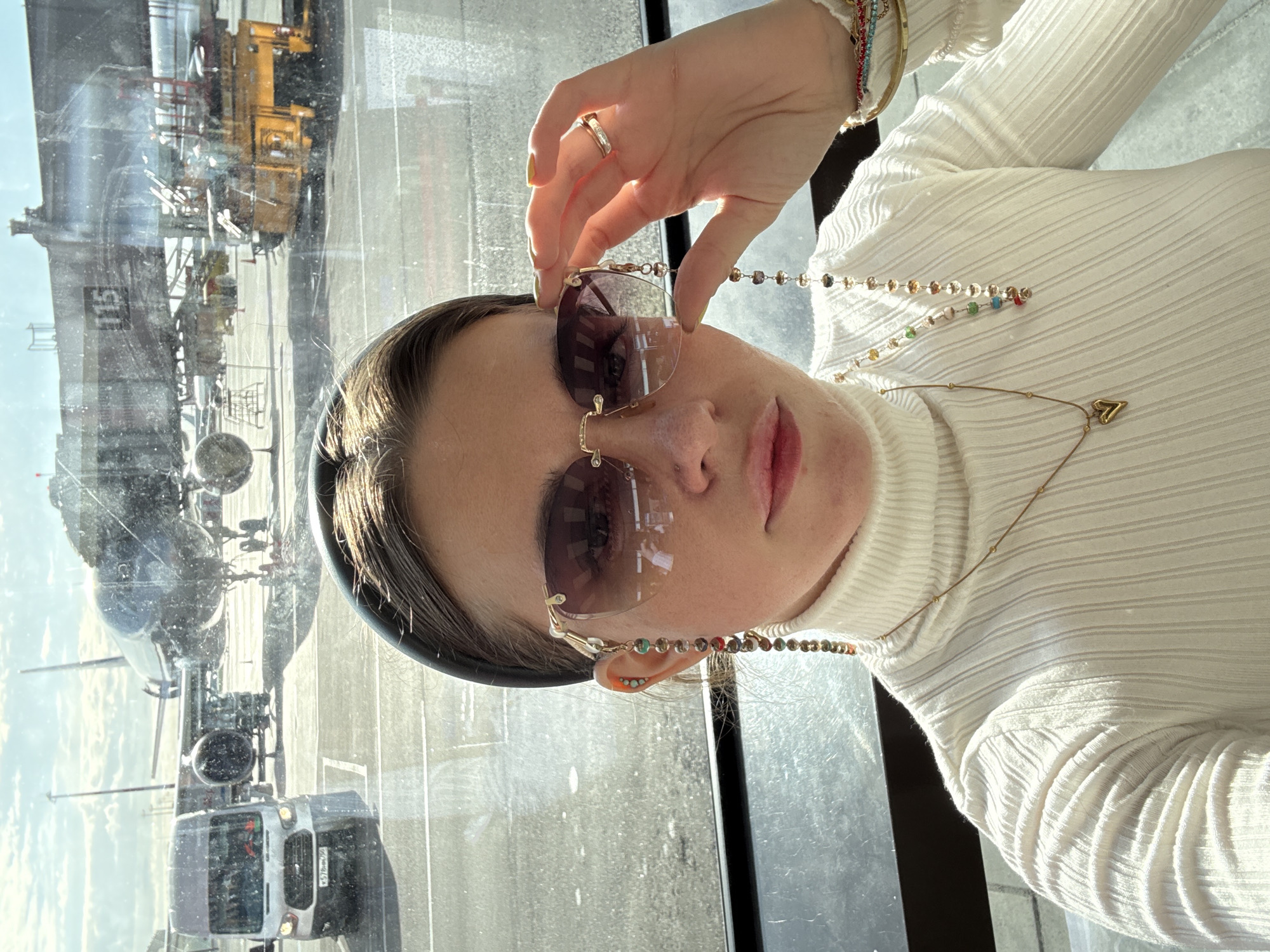 A person is taking a selfie in front of a window at what appears to be an airport. The subject is wearing a white turtleneck sweater, sunglasses, and jewelry. Through the window, an airplane and vehicles are visible. The airplane has a logo of what is likely Russian Airlines. The vehicles look like airport service vehicles. The location appears to be in Khimki, Russia based on the airplane logo in the reflection.