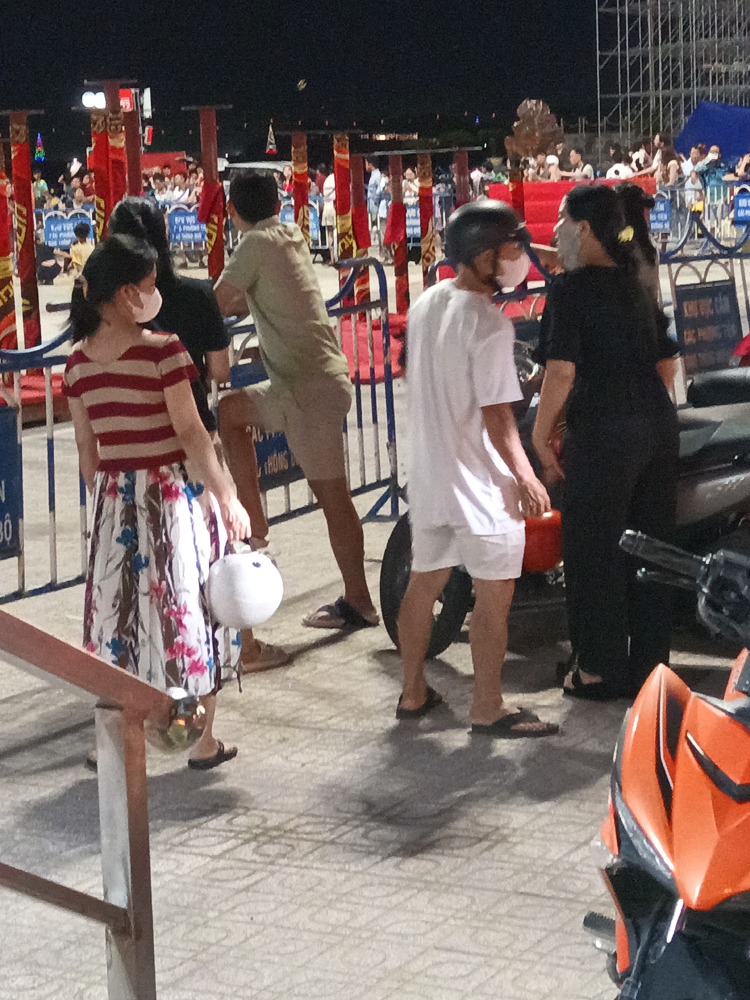 The image depicts a nighttime scene in Nha Trang, Vietnam, likely at a public event or market, given the presence of numerous people, temporary structures, and festive lighting. In the foreground, a young woman with dark hair is wearing a red and white striped t-shirt and a long, floral-patterned skirt. She is holding a white helmet in her right hand and appears to be walking. Beside her, a man in a light green t-shirt and beige shorts is facing away from the camera, looking towards a crowd. To the right of these two individuals, a person wearing a white t-shirt and white shorts, with a black helmet on their head, is facing towards the woman. Behind them, another person in a black outfit is visible. In the background, a crowd of people are gathered, with red and gold decorative structures and scaffolding suggesting a stage or event setup. The ground is paved with patterned tiles, and there are several motorcycles visible, including a prominent orange one in the lower right corner. The lighting suggests artificial illumination from overhead sources and possibly decorative lights.