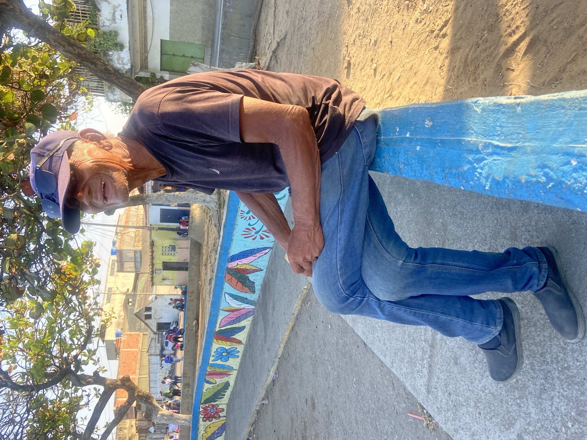 An elderly man wearing a dark blue t-shirt, blue jeans, and a blue baseball cap is sitting on a painted blue concrete structure. He is smiling. The structure is decorated with painted flowers and leaves. Behind the structure, there is a partially obscured view of buildings and people, typical of urban scenes in Venezuela. The image suggests the location is Higuerote, Venezuela. A tree with green leaves is visible on the left side of the frame. The background is blurry with sunlight.