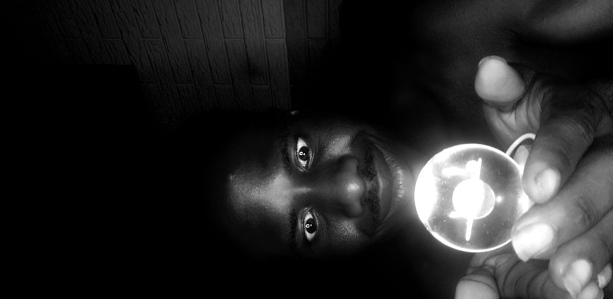 A monochrome, low-angle close-up photo shows a person with dark skin in a dark room holding a small, circular object illuminated from behind. The person is facing the camera with a slight smile. Their fingers are visible around the object, which has a pattern of lines and a circle within it. The background includes a textured wall partially visible in the upper left. Given the information about Narayi, Nigeria, this scene is likely set indoors in a private residence in that location. The photograph suggests a personal moment or artistic expression rather than a typical street scene.