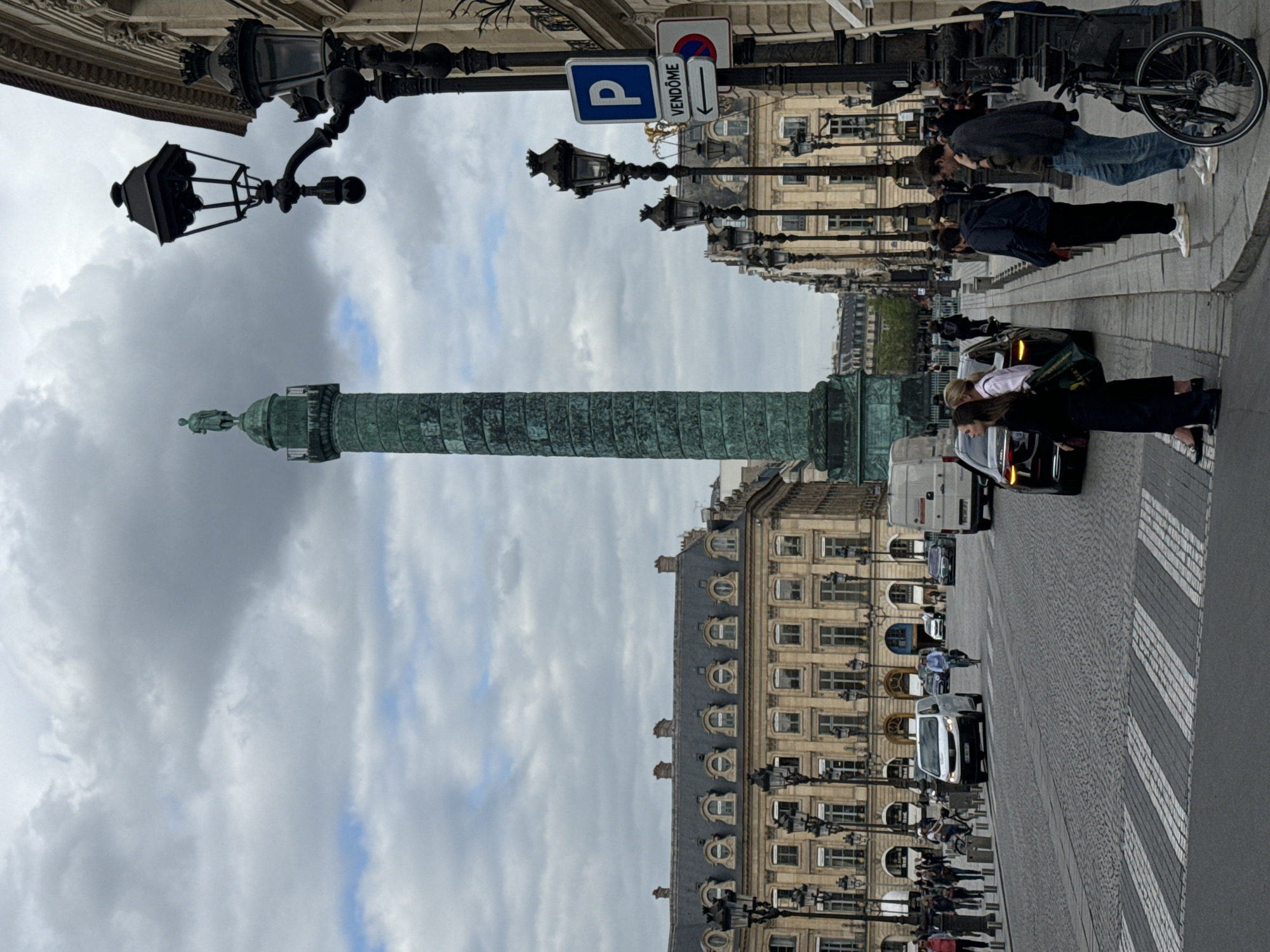 The image captures the Place Vendôme in Paris, France, featuring the Vendôme Column prominently in the center. The sky above is cloudy. On the right side of the image, a building with multiple windows and arched details is visible. A row of black lampposts lines the sidewalk in front of the building. Several people are present, some standing on the sidewalk, others crossing the street at a pedestrian crossing. Cars and other vehicles are moving along the road. A parking sign with the word "Vendôme" and a directional arrow is affixed to a lamppost. A bicycle is parked on the sidewalk near a group of people.