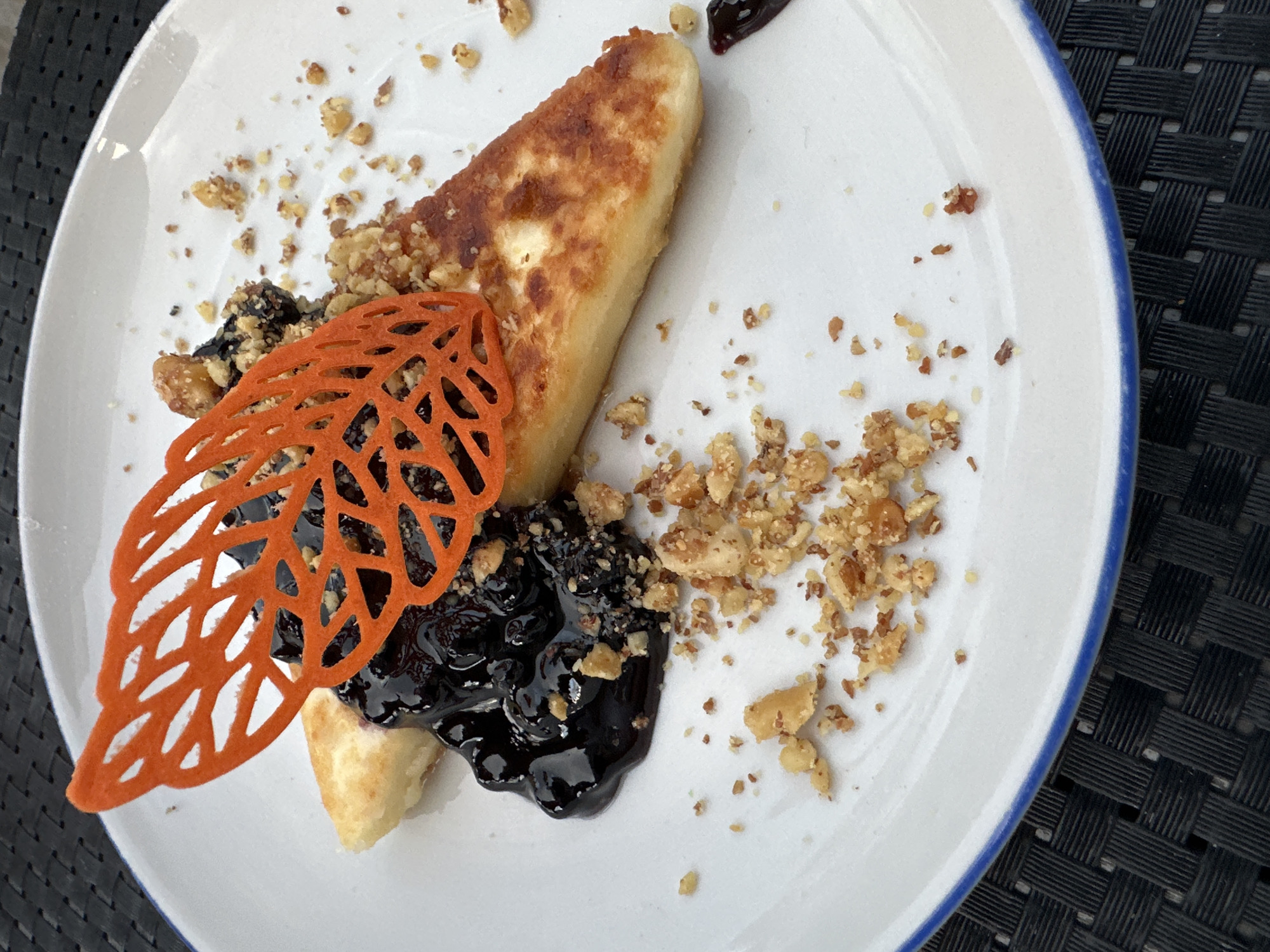 A close-up, overhead shot displays a dessert on a white, circular plate with a blue rim, positioned on a black, woven placemat. The dessert consists of a wedge of fried cheese, topped with a dark, glistening berry compote and garnished with chopped nuts. An intricate, orange lace-like decoration, resembling a leaf or feather, is placed on top of the compote. Crumbs of nuts are scattered across the plate and around the dessert. The lighting suggests it is daytime, possibly outdoors given the warm tones. The exact location in Enina, Bulgaria, is not discernible from the image.