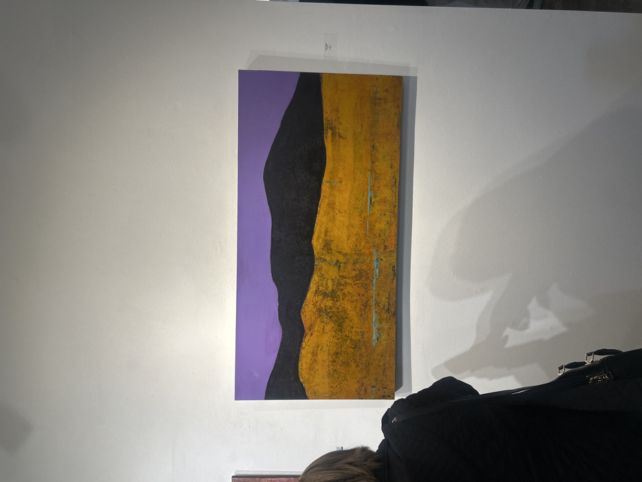 A vertical abstract painting hangs on a white wall. The painting features a solid lavender section to the left, adjoined by a jagged black shape. To the right of the black shape is a textured gold area with vertical teal lines. Below the painting are partial views of a dark jacket, a light-haired person, and a black case. A faint shadow is cast on the wall above the jacket.