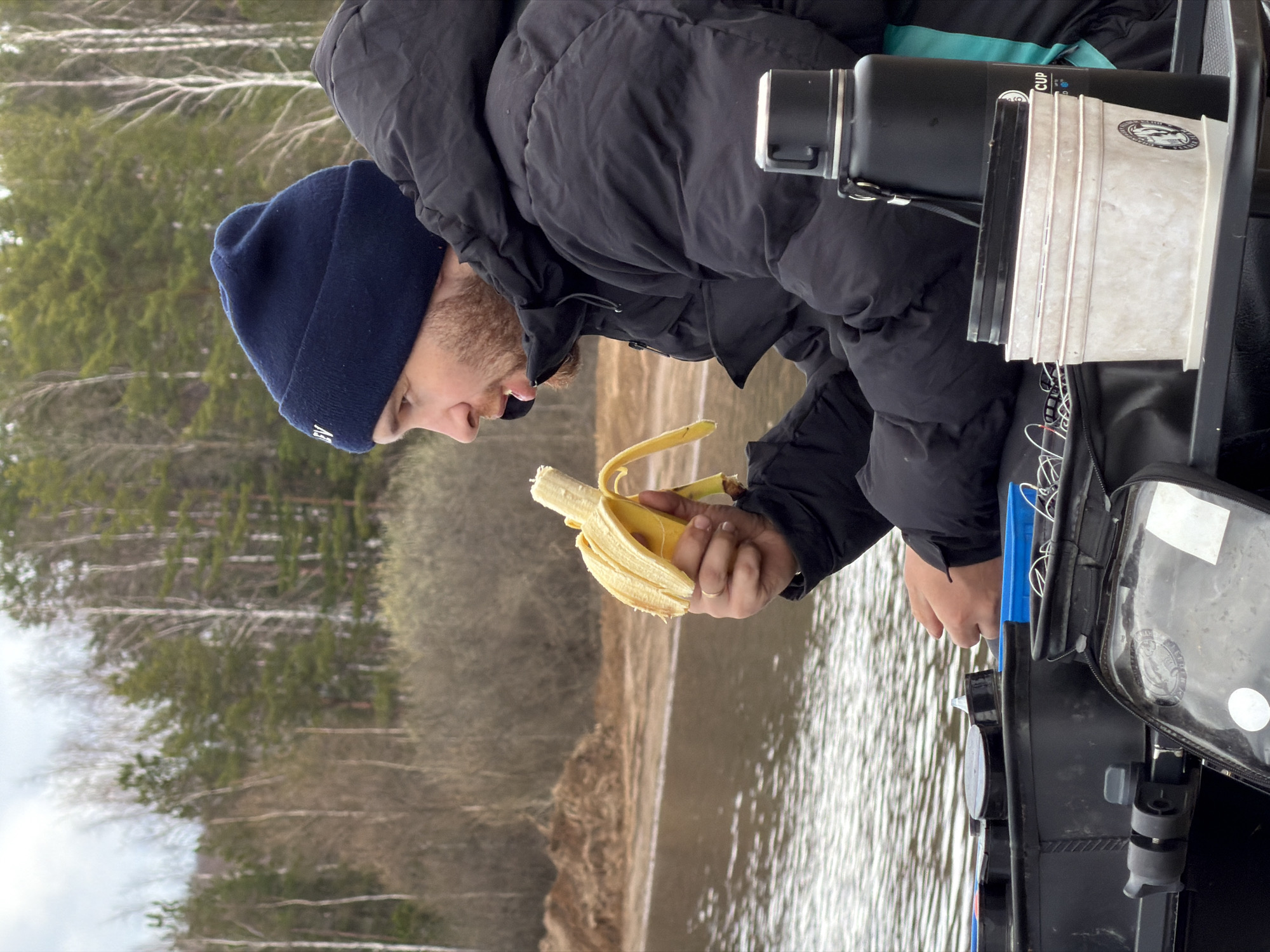 A man in a black puffer jacket and a navy blue beanie is peeling a banana near a body of water. The background shows a shoreline with trees, possibly indicating a lake or river. The man is positioned next to a black container with various items attached, including a water bottle and a clear plastic document holder. The location is suggested to be near Chaikovsky, Russia, based on the request. The man appears to be preparing to eat the banana. The water is rippled, reflecting the overcast sky.
