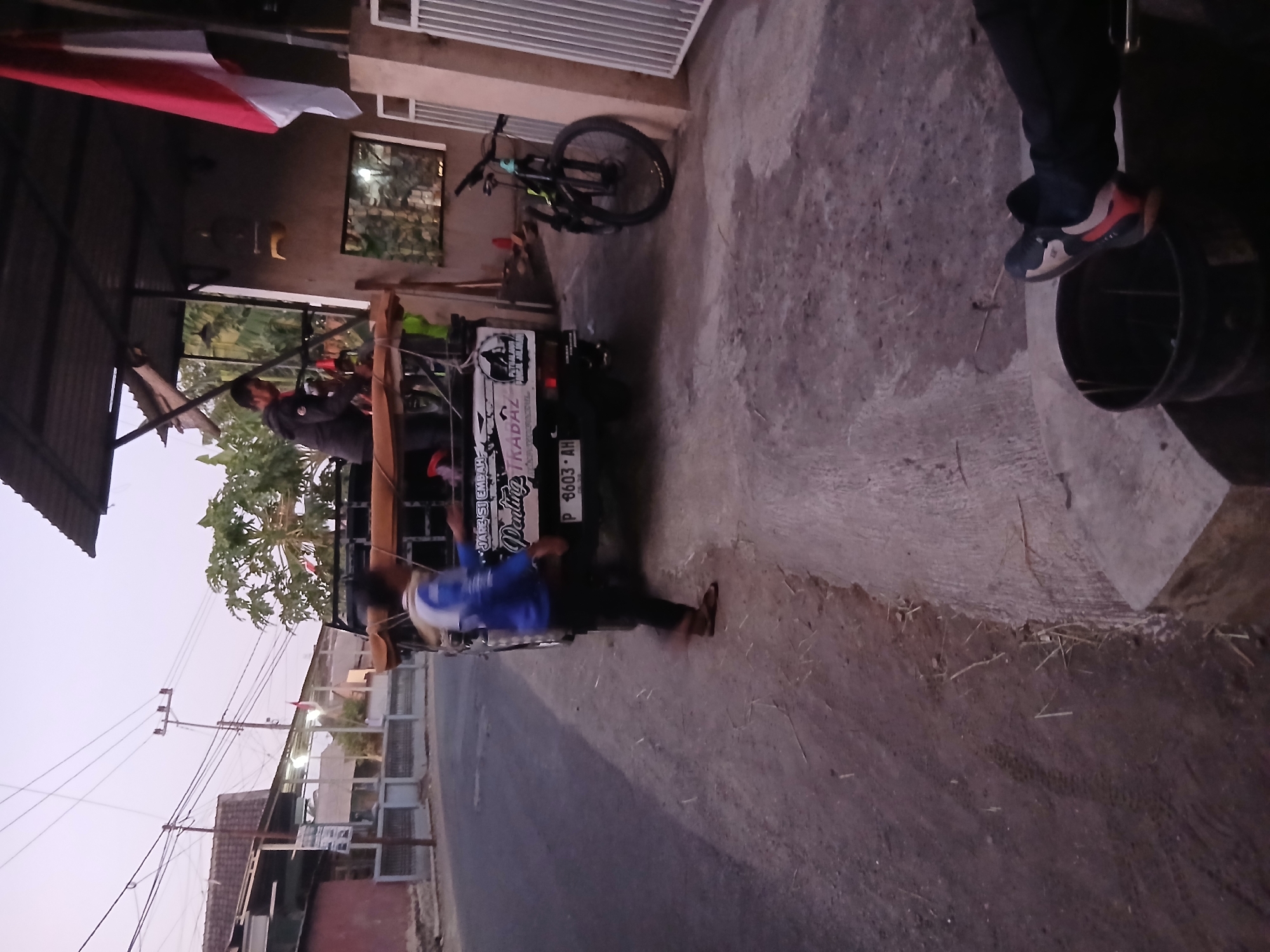 The image depicts a street scene in Kademangan Kulon, Indonesia, at dusk. A small pickup truck, loaded with wooden planks and covered with a brown tarp, is parked on the side of a paved road. Two individuals are near the truck. One, wearing a blue jacket and dark pants, is partially visible and appears to be walking away from the truck. The other individual, in a dark jacket, is standing on the truck bed, seemingly engaged in an activity. A bicycle with a black frame and green accents is propped up against a wall to the right of the truck. The environment includes a building with a corrugated roof structure and an Indonesian flag prominently displayed. The ground is a mix of dirt and pavement. In the foreground, a person's legs and feet, wearing athletic shoes, are visible near a black bucket. The lighting suggests the end of the day.