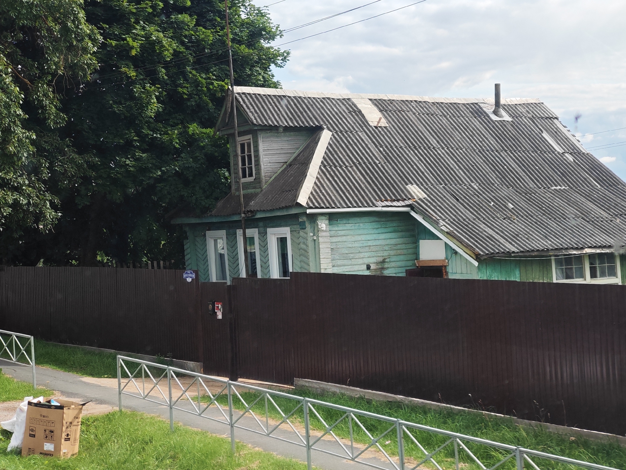 The image depicts a two-story wooden house with peeling green paint and a corrugated metal roof. The house is partially obscured by a tall, dark brown metal fence. To the left of the house, a dense canopy of green trees provides shade. In the foreground, a paved pathway runs alongside a metal railing, with patches of green grass and a discarded cardboard box. The sky above is overcast with scattered clouds. A street sign with the inscription "Советская" (Sovetskaya) and the number "15" is visible on the fence, indicating the location is Sovetskaya Street, number 15, in the city of Выползово, Russia.