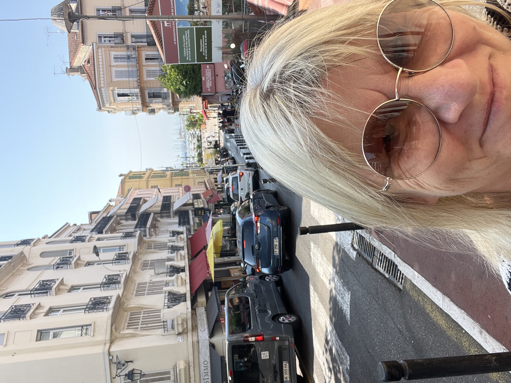 In Cannes, France, a woman with blonde hair and sunglasses takes a selfie in front of a street lined with multi-story buildings. The street is busy with traffic, including a grey van and a blue hatchback, and has a crosswalk. Some buildings have awnings and balconies, and some appear to be shops. In the background, boats are visible in the water. There are streetlights and signs, one in particular reading "LE RENOUVEAU DU JARDIN DES SAVEURS ET DES ODEURS SUR LE TOIT DU MARCHÉ FORVILLE" and another indicating "RÉNOVATION ET EMBELLISSEMENT DE LA HALLE ET DE SES ABORDS". The sky is clear and blue.