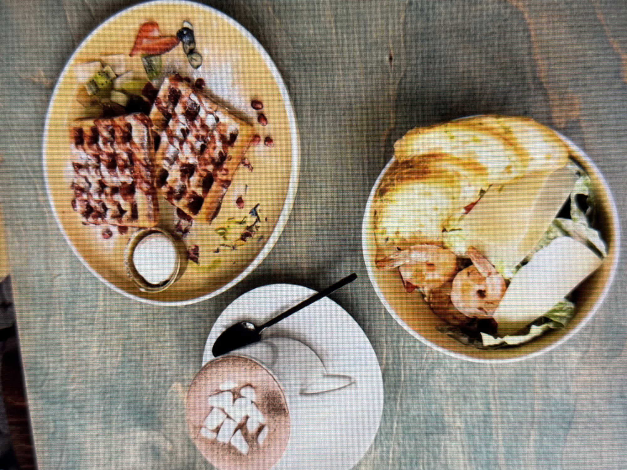 A top-down, high-angle shot captures a rustic wooden table with three food items in Sirius, Russia. To the left, a white mug filled with hot chocolate and marshmallows sits on a white saucer, accompanied by a black spoon. In the center, a yellow plate holds two square waffles drizzled with chocolate syrup and dusted with powdered sugar, garnished with pomegranate seeds, kiwi slices, and a strawberry heart. To the right, a white bowl contains a salad with shrimp, lettuce, and what appears to be cheese and bread. The overall scene suggests a casual meal or dessert setting.
