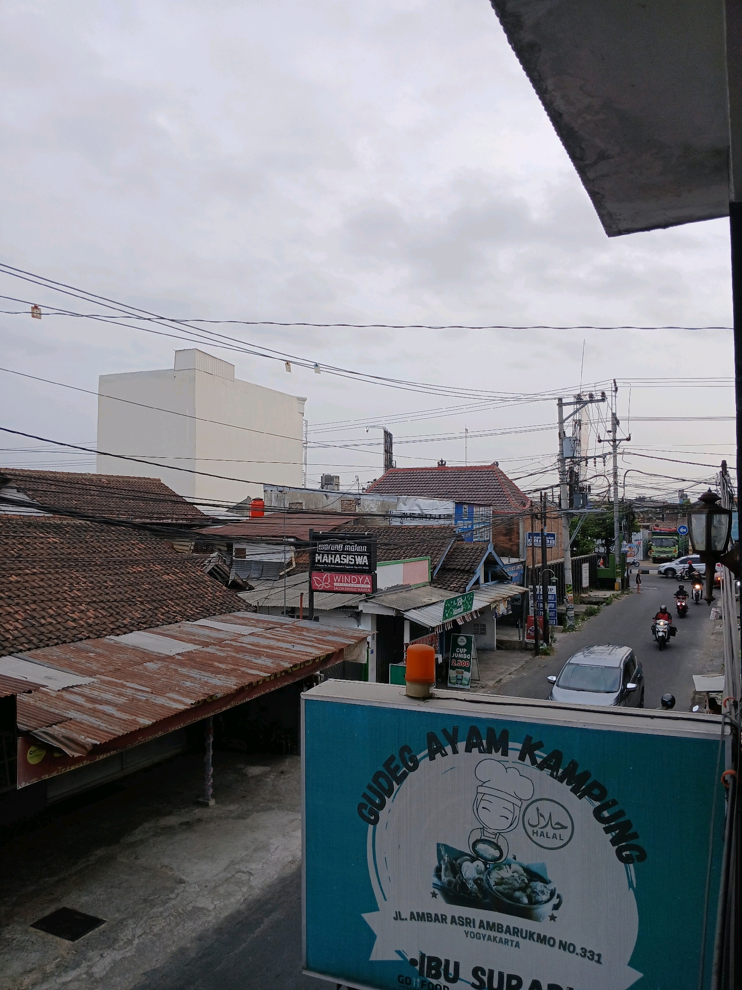This is an outdoor, eye-level shot of a street scene in Depok, Indonesia. The sky is overcast and a light grey color. In the foreground, there is a large, turquoise billboard advertising "GUDEG AYAM KAMPUNG" with a cartoon chef and a "HALAL" symbol. Below this is text that reads "JL. AMBAR ASRI AMBARUKMO NO.331 YOGYAKARTA" and "IBU SUPARDI". To the left of the billboard is a weathered, rusted tin roof extending over what appears to be a shop or building.
Further into the scene, several buildings with tiled roofs are visible. One building stands out with its stark white, multi-story facade. A prominent network of electrical wires and poles crisscrosses the sky and street. The street itself is paved and shows signs of activity. Two motorcycles are seen traveling down the road, one with a rider wearing a red jacket, and another with a rider wearing a black jacket. A silver sedan is parked on the side of the road, and a green truck is visible further down the street. Various shop signs are present, including one for "WARUNG MAKAN MAHASISWA" and "WINDYA SALON KECANTIKAN WANITA". The overall impression is a busy urban environment with a mix of residential and commercial structures under a cloudy sky.