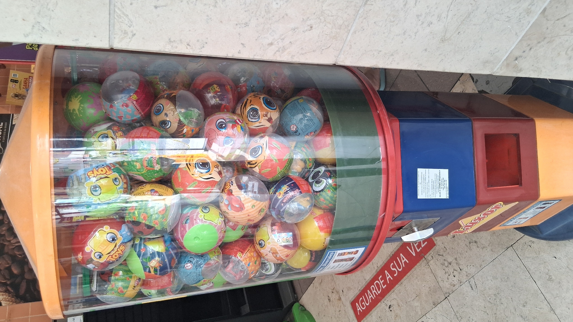 The image shows a bright, colorful gumball or toy dispenser machine filled with numerous small, individually wrapped plastic balls. These balls are adorned with various cartoonish characters, including animals like tigers and monkeys, and have vibrant colors and playful designs. Some balls have clear plastic wrappers that allow their inner colors and designs to be seen. The dispenser itself is made of clear plastic, revealing the densely packed contents, and has a red rim. The exterior of the machine is a combination of orange, blue, and red plastic, with some sections appearing to be darker or a different material.
A notable detail on the machine is a red sticker with white text that reads "AGUARDE A SUA VEZ," which translates from Portuguese to "WAIT YOUR TURN." There are also other labels and text elements visible on the machine, including "Toys" and what appears to be a company name, "SILVA & CARMO DIVERSÕES." The machine is positioned in an indoor setting, likely a public space, as suggested by the tiled floor and a hint of what might be a store or commercial area in the background with some packaging visible. The lighting appears to be artificial and even, suggesting it's likely indoors during daylight hours. The overall impression is of a fun, engaging dispenser designed for children or as a novelty item.