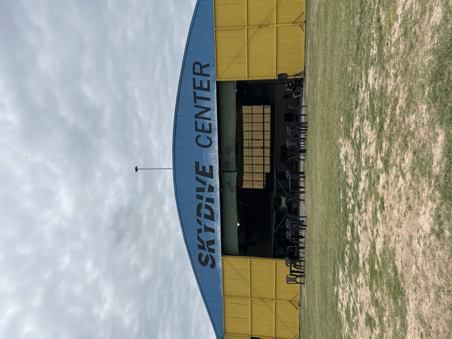The image displays the exterior of an arched hangar building labeled "SKYDIVE CENTER" in dark lettering on its blue roof, located at an aerodrome in Chascomús, Argentina. The hangar features large yellow corrugated metal sliding doors, which are partially open, revealing a light aircraft inside. In front of the hangar, on a sparse grassy area with visible soil, approximately five black plastic tables are arranged with multiple accompanying black plastic chairs. To the right, near the open hangar section, a black speaker or audio equipment is positioned on the ground. Two individuals are partially visible inside the hangar opening on the right, near the speaker. The sky above is overcast with light grey clouds. A thin, dark pole extends vertically from the hangar roofline into the sky, where a small, dark, unidentified object is faintly visible.