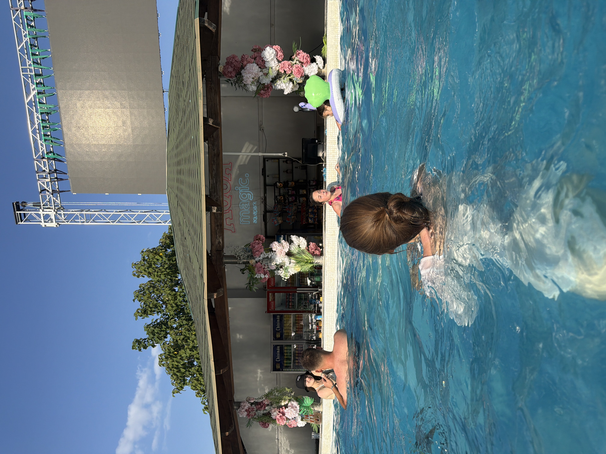 The image depicts a sunny day at an outdoor aquapark in Sociteni, Moldova. Several people are enjoying the water. In the foreground, a person with brown hair tied in a bun is submerged in the blue pool, with part of their arm and a white garment visible. To their right, another person's head and shoulders are visible above the water. Further in the background, a young girl with a green inflatable ring floats near the edge of the pool. Beside the pool, a shaded structure with a sloped roof houses a bar or shop with refrigerators stocked with drinks and shelves displaying various items. Floral decorations, with pink and white flowers, are placed near the edge of the pool and on the structure. In the distance, a large screen is mounted on a metal truss structure. A tree with green foliage is visible against a clear blue sky with a few wispy white clouds. The overall atmosphere appears relaxed and recreational, suggesting a leisure activity on a warm day. The text "magic. aquapark" is visible on the wall of the structure.