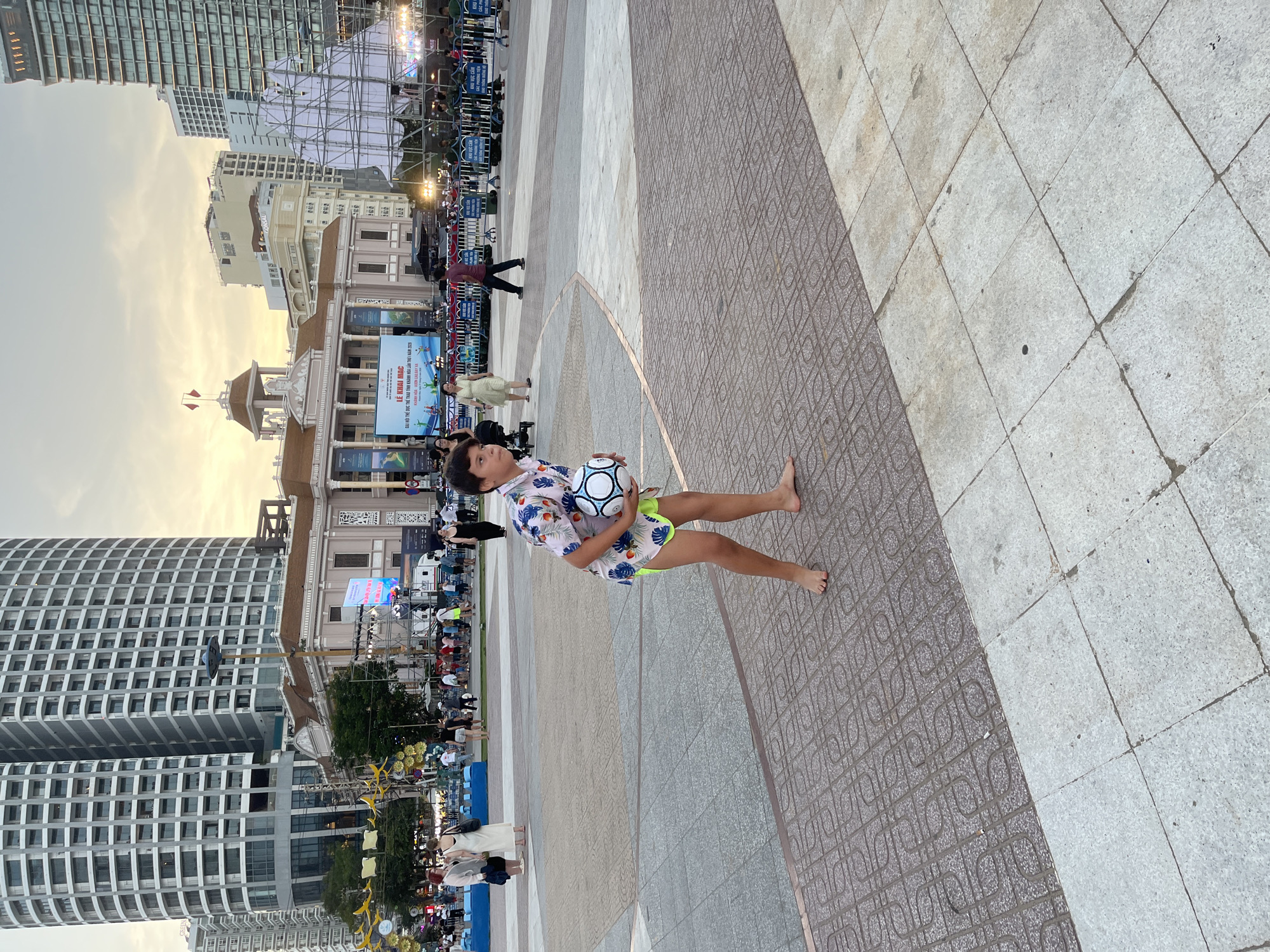 A young boy, barefoot and wearing a floral shirt and neon green shorts, stands in the foreground holding a black and white soccer ball. He is in an outdoor public plaza with a patterned tile floor. In the background, a crowd of people are gathered, and several large buildings, including a multi-story cylindrical building with many windows, are visible. There is also a stage or setup with scaffolding and a large screen displaying text. The sky is overcast and appears to be dusk. The scene is set in Nha Trang, Vietnam, likely in a central area such as a public square or promenade.