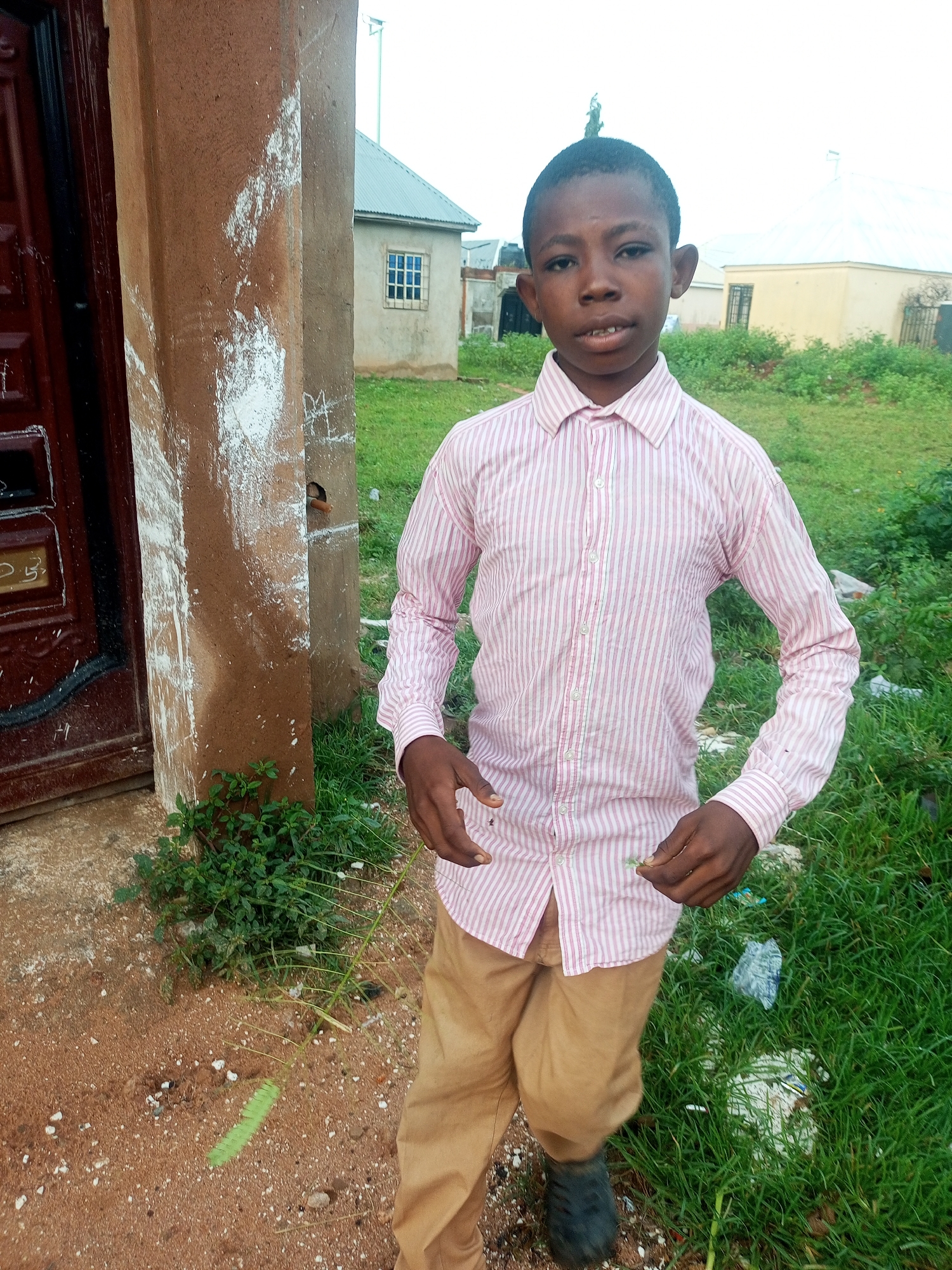 A young boy stands on a dirt path beside a plastered, brown pillar with white paint splatters and a dark red door. He wears a pink and white striped button-down shirt and tan pants. In the background, a building with a gray, corrugated metal roof and a small window is visible, along with a yellow building. The ground is covered in patchy green grass and scattered debris.